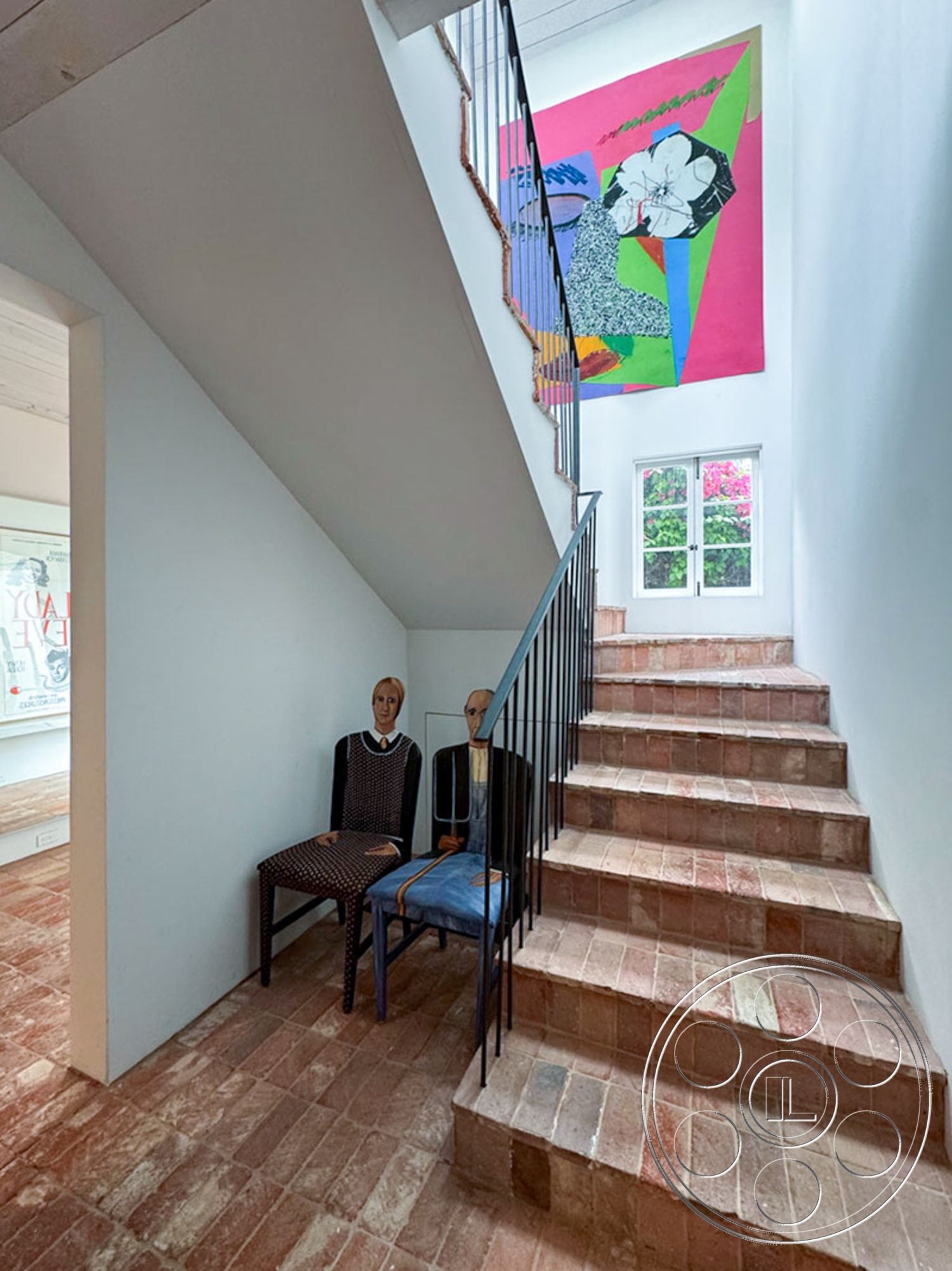 Spanish 165 - artwork on wall, natural light from window, textured plaster wall finish, white painted walls, exposed brick accent wall, metal stair railings, interior brick staircase steps, open staircase design, vintage chairs, faux mannequin decorations, rectangular staircase layout, warm-toned brick flooring