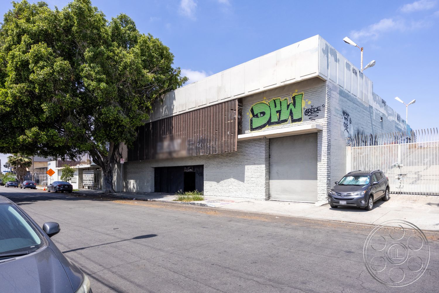 Warehouse 61 - urban setting, large industrial windows, nearby palm trees, graffiti wall art, single-story building, concrete sidewalk, metal roof details, exterior brick walls, exposed concrete façade, metal slat screening, street-facing commercial structure, drainage gutters, landscaped tree borders, overhead exterior lighting, modern industrial architecture, facade with multiple textures
