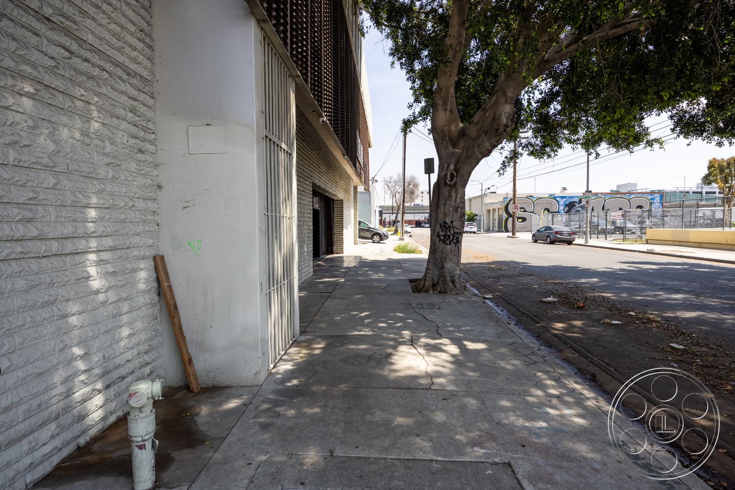 Warehouse 61 - urban environment, unmaintained landscaping, industrial architectural style, surrounding city view, street-level commercial space, exterior wall treatments, textured concrete block walls, white painted concrete wall, sidewalk concrete surface, rough concrete pavement, urban streetscape, tree-lined sidewalk, metal grating facade details, utility access points, ashphalt street, vintage street elements