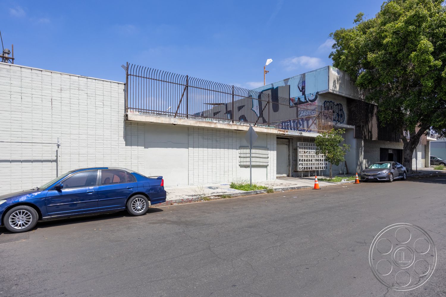 Warehouse 61 - urban setting, exterior concrete patio floor, flat roof structure, overhanging roof eaves, exterior concrete block walls, exterior graffiti accent wall, metal security fencing, street-side parking area, paved asphalt road, taxiway tree landscaping