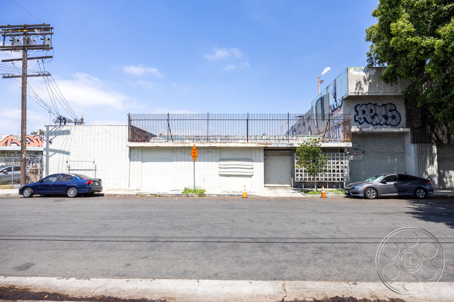 Warehouse 61 - urban setting, street view, graffiti wall art, open parking spaces, flat roof design, exterior white painted walls, minimalist architectural style, rectangular building shape, asphalt street surface, exterior concrete block walls, metal security fence, surrounding power lines, landscaped tree area, commercial building façade, hardscape pavement, city landscape