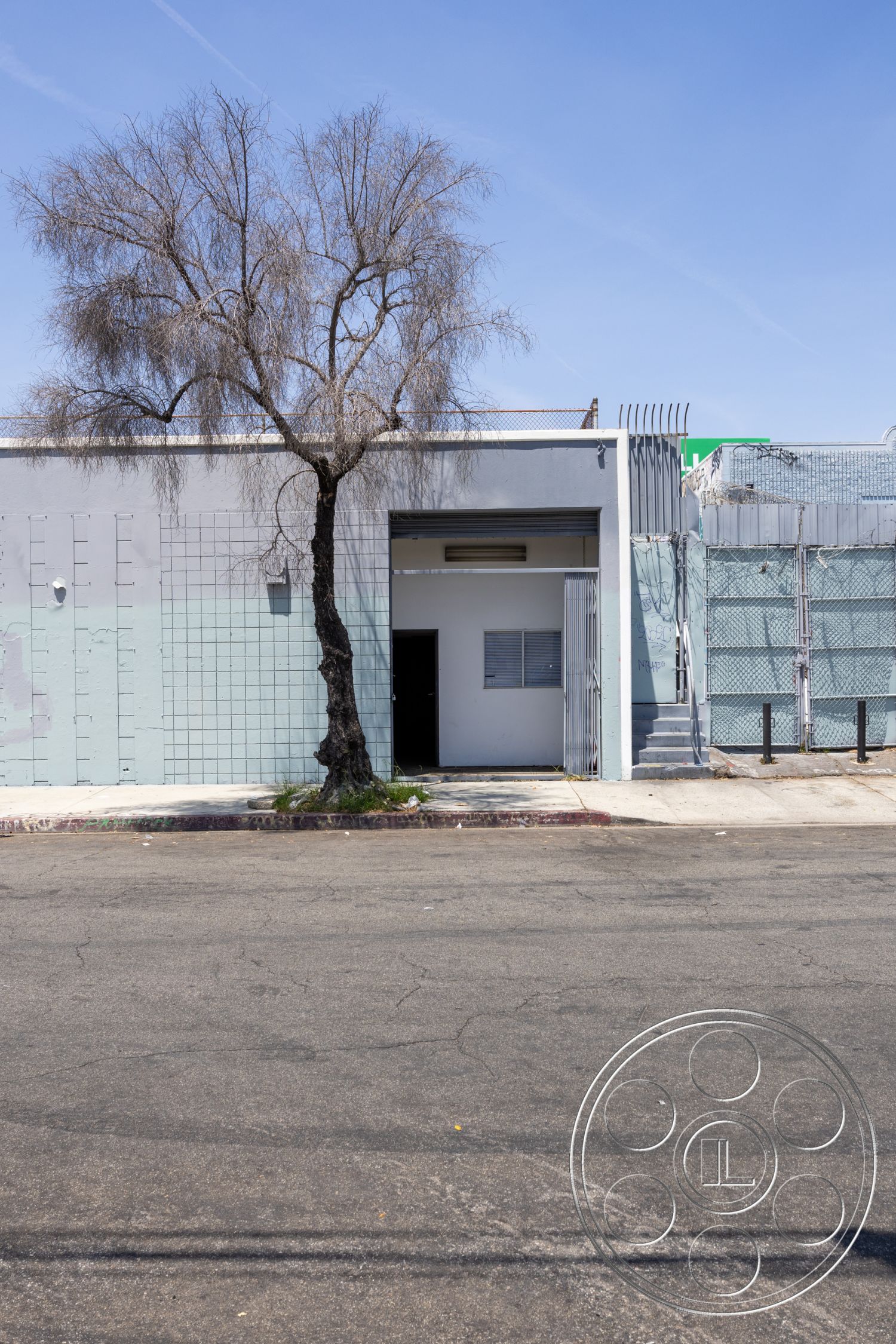 Warehouse 61 - city street view, concrete sidewalk, industrial environment, flat roof design, minimalist architectural style, steel railing staircase, urban commercial building exterior, exposed concrete surface, metal security fence, gray painted concrete walls, simple rectangular doorway, partial brick accent wall, lonely leafless tree in urban setting, hard flat street pavement, plain window design