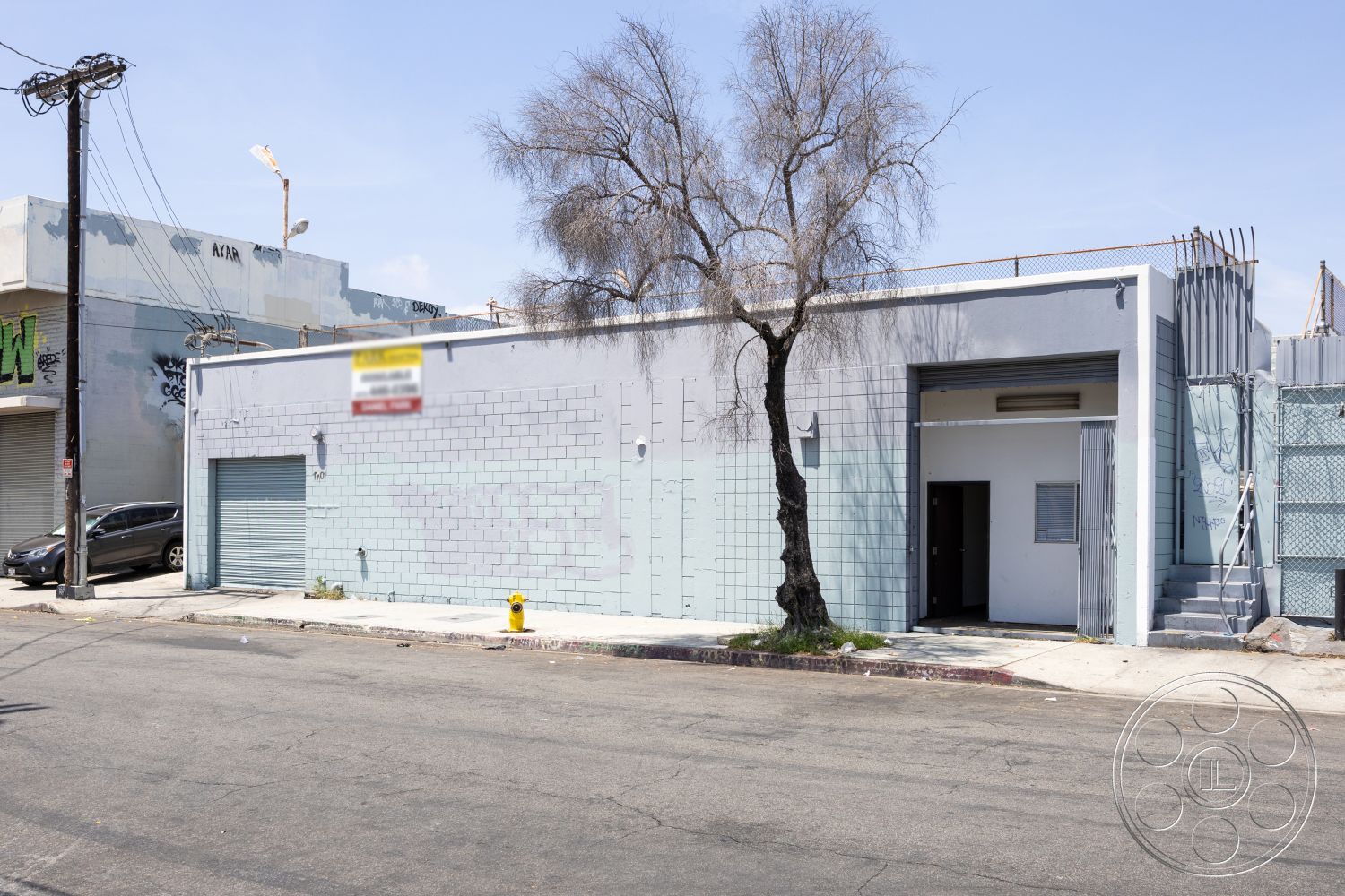 Warehouse 61 - city street view, concrete sidewalk, flat roof design, minimalist architectural details, single-story commercial structure, exterior concrete ramp, metal utility pole, exterior concrete block walls, exterior gray painted concrete walls, industrial-style loading dock door, metal security grilles on windows, asphalt street setting, urban industrial environment, concrete entry steps, surrounding street tree, plain metal door entry, faded exterior wall paint, curved shape of loading dock