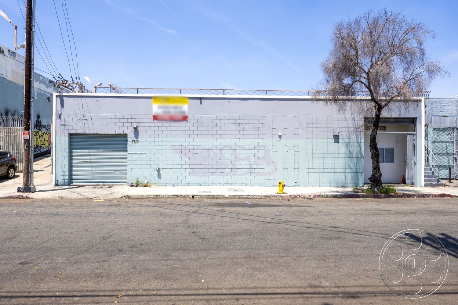 Warehouse 61 - asphalt driveway, single-story building, tree in front yard, concrete sidewalk, overhead power lines, urban setting with street view, metal security gate, commercial building facade, exterior concrete block walls, gray painted exterior, metal roll-up garage door, minimalist industrial architecture, fenced outdoor area, surrounding graffiti art