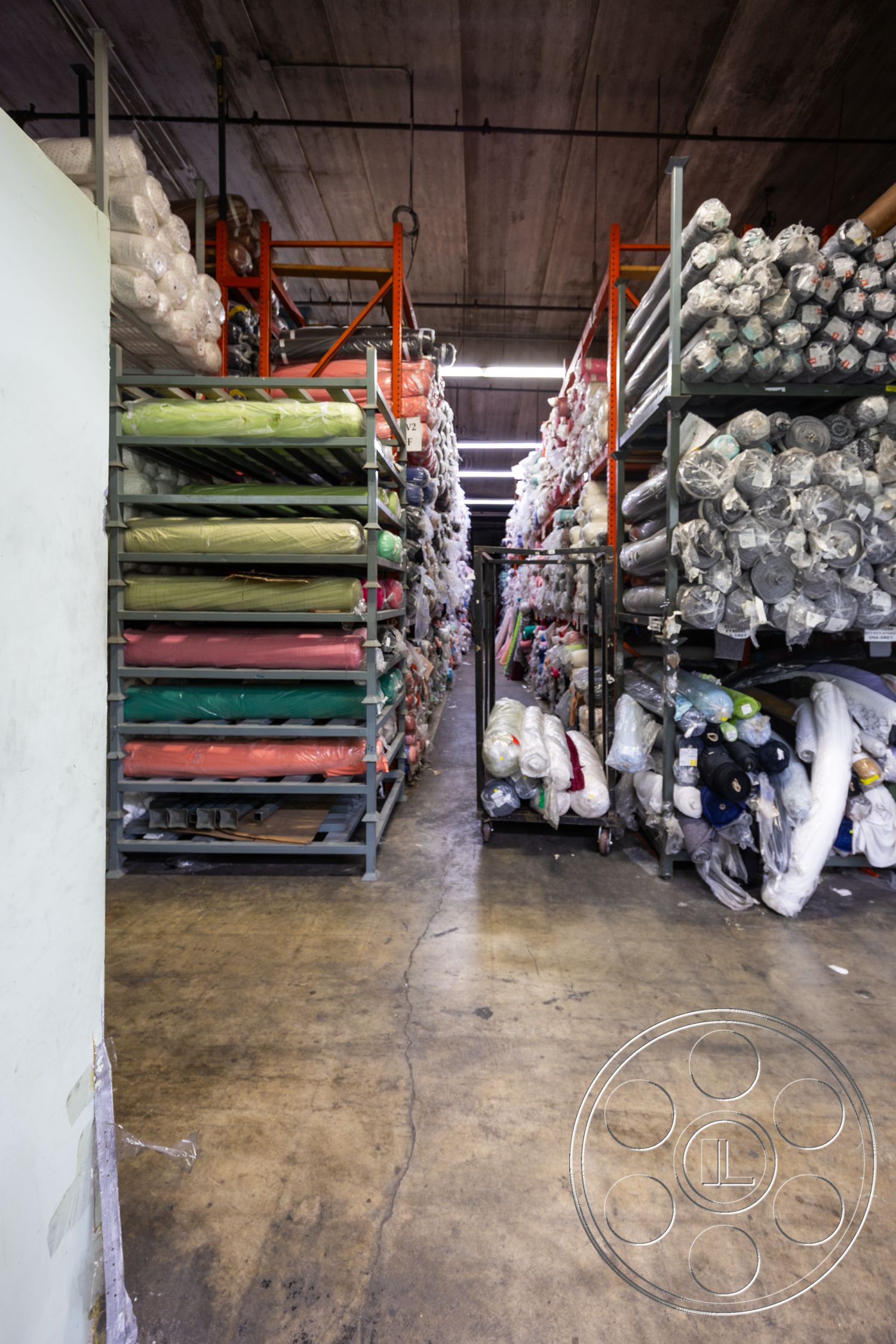Warehouse 61 - metal shelving units, exposed concrete floor, concrete wall finishes, rolls of fabric storage, industrial warehouse interior, stacked textile materials, high ceilings with exposed beams, fluorescent overhead lighting, narrow aisle layout, warehouse storage area