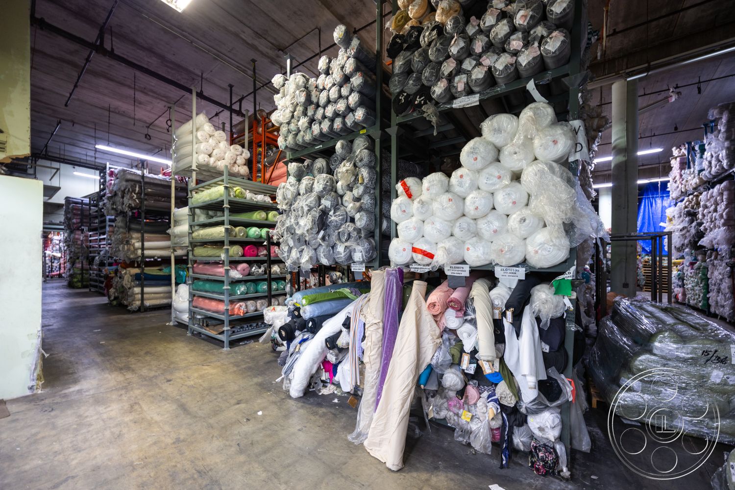 Warehouse 61 - neutral wall colors, industrial lighting fixtures, interior fabric storage area, open shelving for textiles, concrete floor in fabric warehouse, plastic wrapped fabric rolls, colorful fabric spools, stacked bolt fabrics, metal shelving units for storage, wide aisles for material accessibility, textile retail environment, large open warehouse space, organized fabric display, floor-to-ceiling fabric inventory, rolls of cotton fabric, polyester fabric storage, nylon fabric rolls, fabric warehouse with abundant shelving, textile market setting, multicolored cotton rolls, fabric bolts with labeled tags, tightly packed fabric stacks, industrial style storage facility