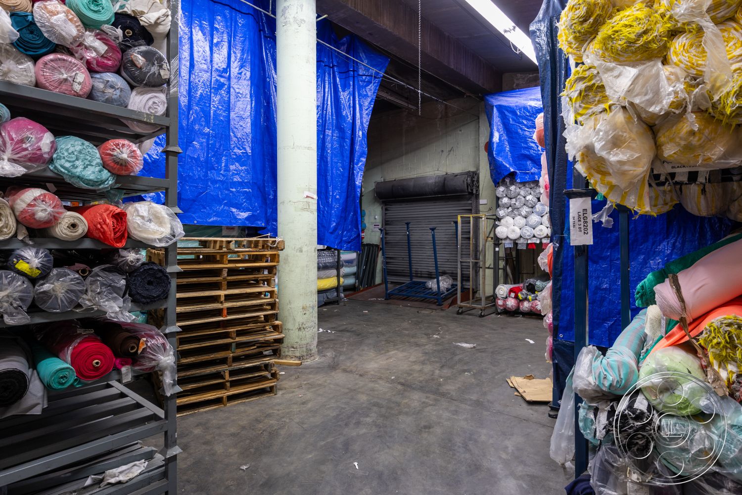 Warehouse 61 - exposed concrete floor, steel support columns, industrial shelving units, textured blue tarpaulin walls, metal pallet storage, wrapped fabric rolls, utility space interior, suspended ceiling grid, open warehouse layout, natural light from overhead fluorescent fixtures