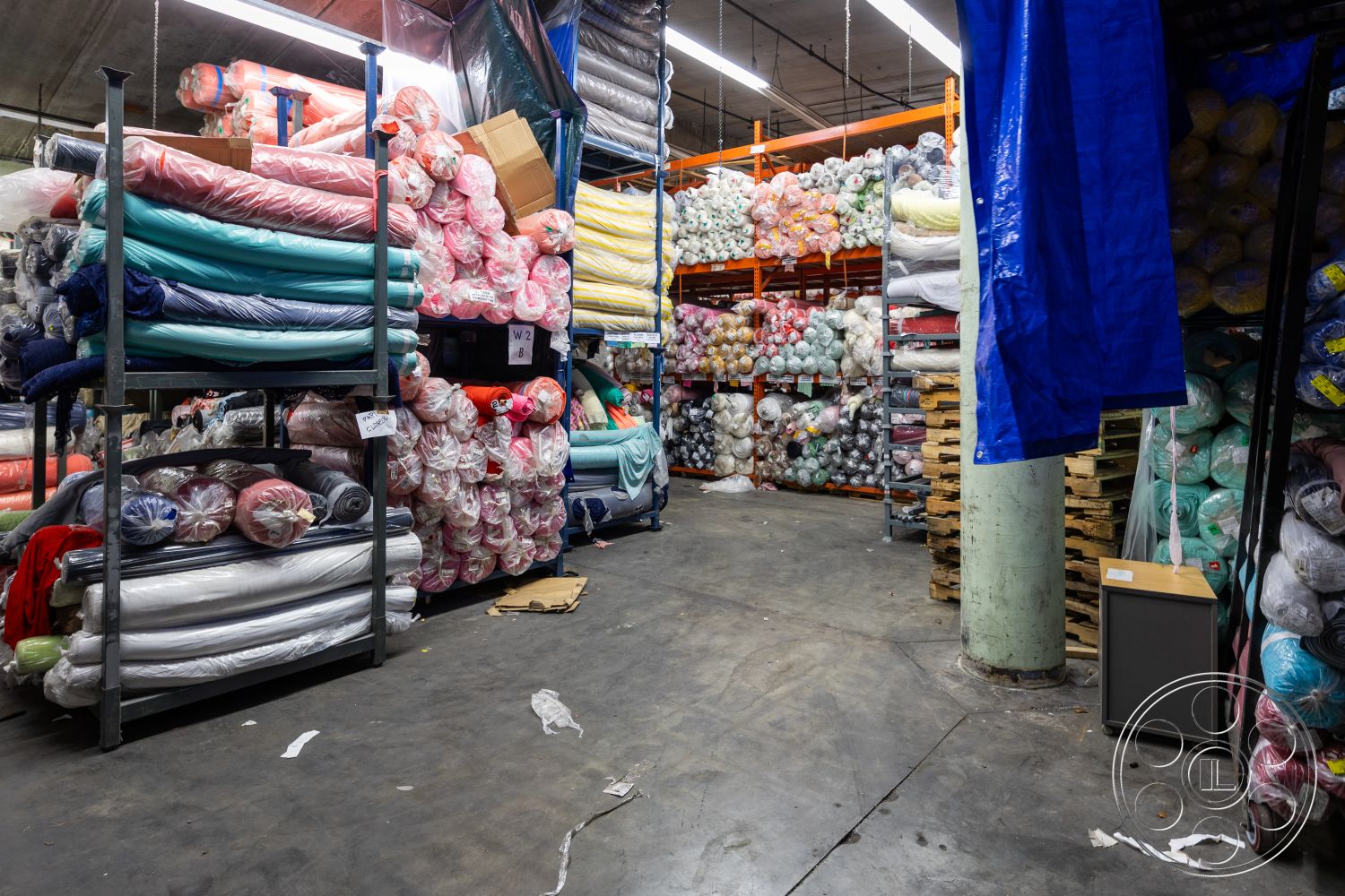 Warehouse 61 - open floor plan, concrete floor, high ceiling, industrial lighting, organized chaos, interior warehouse space, steel shelving units, colorful fabric rolls, plastic-wrapped textiles, stacked textile inventory, pallets for storage, overhead storage racks, mixed color palette, functional industrial design, textile warehouse, brightly colored fabric, efficient storage solutions