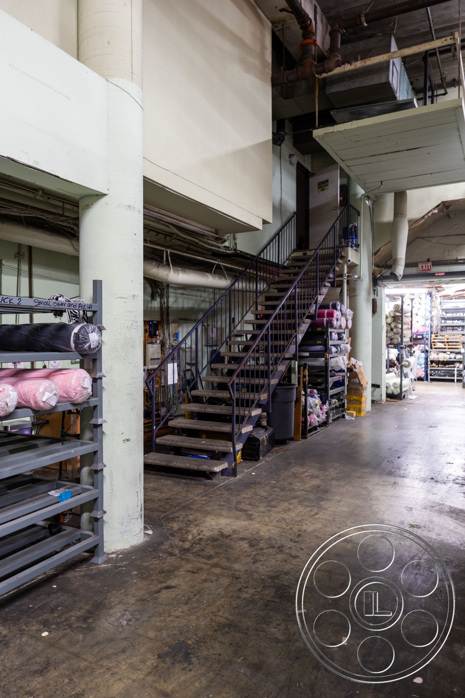 Warehouse 61 - industrial flooring, metal staircase, suspended ceiling panels, exposed concrete columns, functional workspace environment, open warehouse interior, steel railings on staircase, piled fabric rolls storage, concrete floor with stains, overhead ductwork details, stacked storage bins, interior industrial setting, utilitarian design elements, dimly lit warehouse atmosphere, horizontal storage shelves, raw materials display area