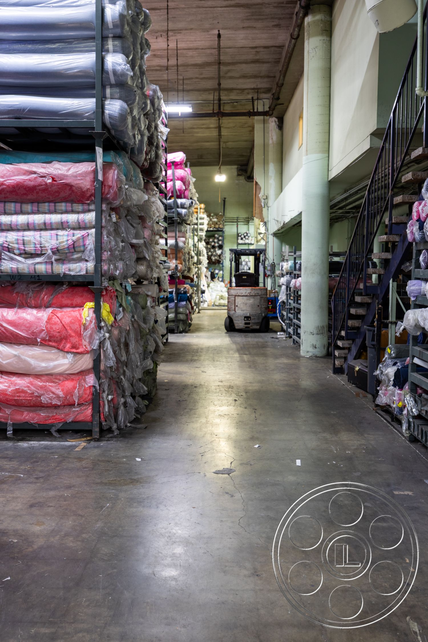 Warehouse 61 - textured concrete walls, metal shelving unit, industrial lighting fixtures, exposed concrete ceiling, commercial space interior, open warehouse layout, concrete warehouse floor, fabric rolls storage area, stacked fabric bundles, steel staircase with metal railings, vinyl flooring in aisles, warehouse storage environment, functional shelving design, goods transportation area