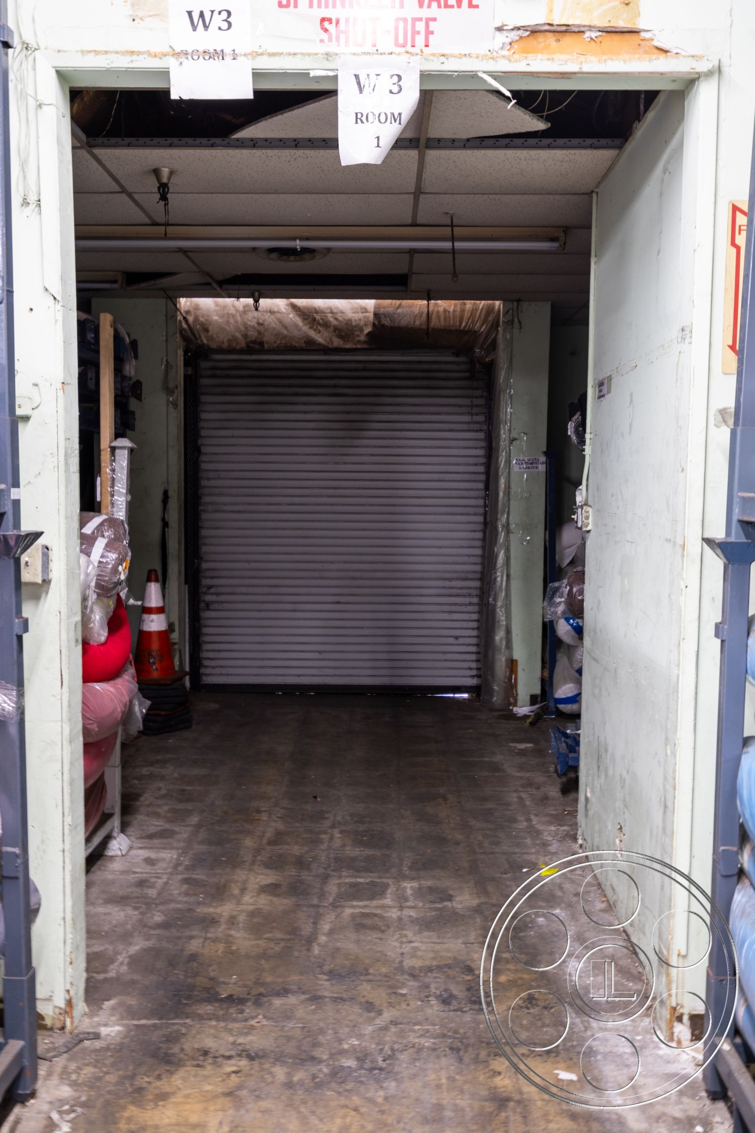 Warehouse 61 - textured concrete surface, painted drywall walls, exposed ceiling tiles, interior storage hallway, vintage concrete flooring, industrial-style ceiling lights, rolling metal security door, backlit signage, sectioned storage area, warehouse-style layout