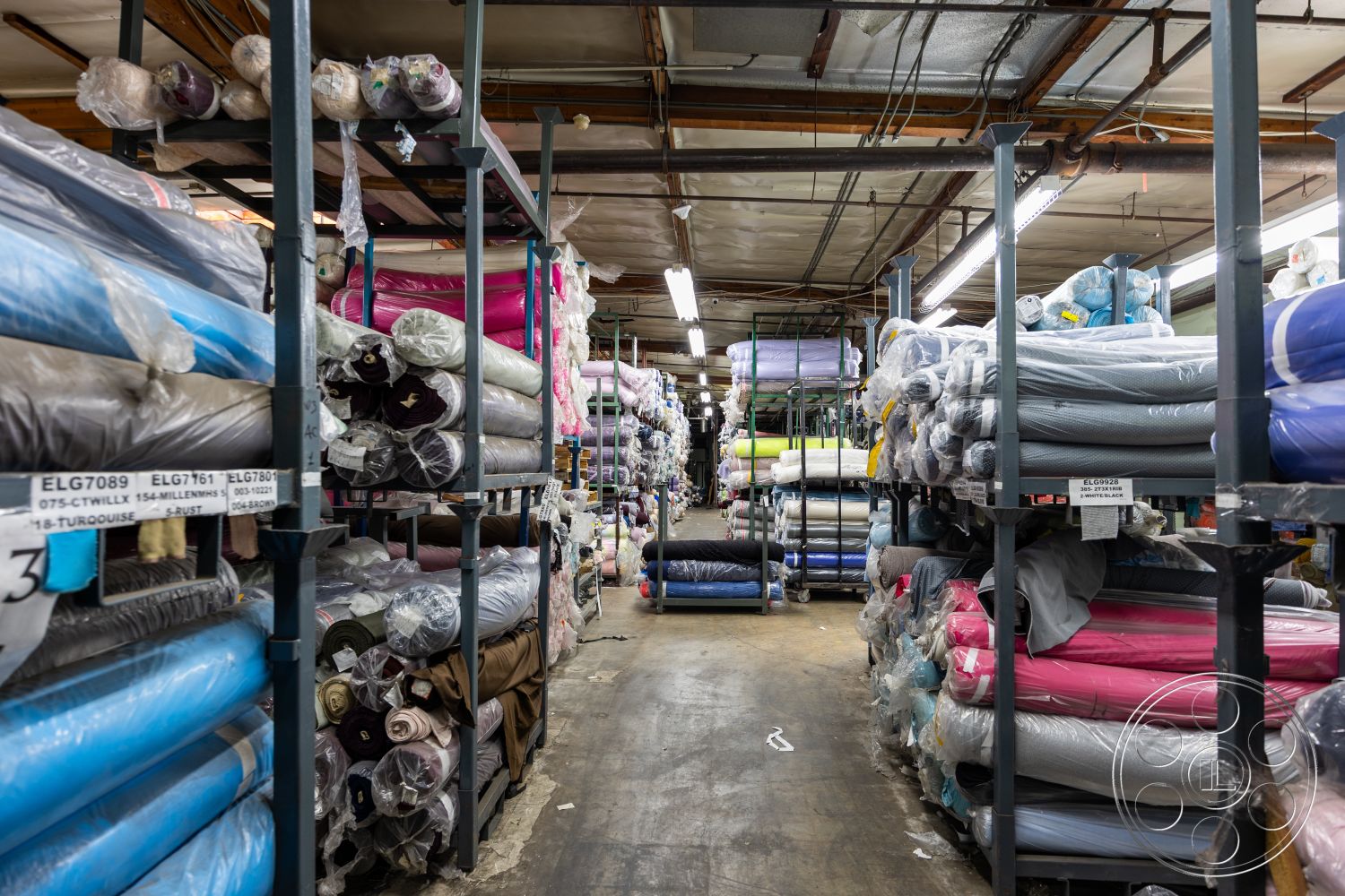 Warehouse 61 - neutral color palette, concrete flooring, functional workspace design, industrial warehouse interior, plastic-wrapped textiles, fabric roll storage area, exposed metal shelving, ambient overhead lighting, rows of fabric rolls, organized inventory space