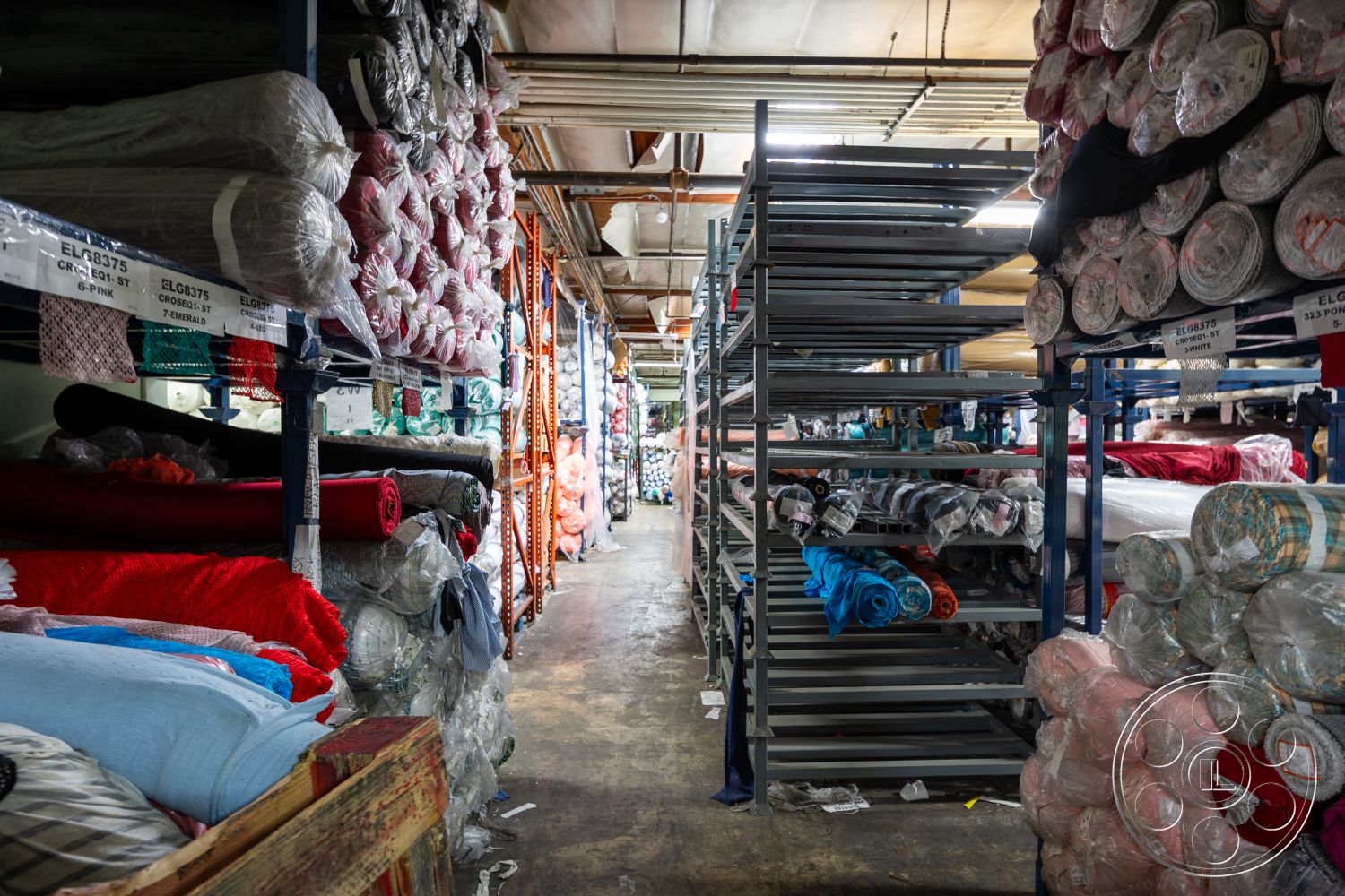Warehouse 61 - concrete floor, overhead fluorescent lighting, industrial warehouse interior, stacked fabric bundles, exposed metal shelving, blue fabric rolls, colorful textile displays, large storage aisle, packed fabric inventory, textile roll storage, organized shelving system, textured fabric surfaces, shelving unit structures