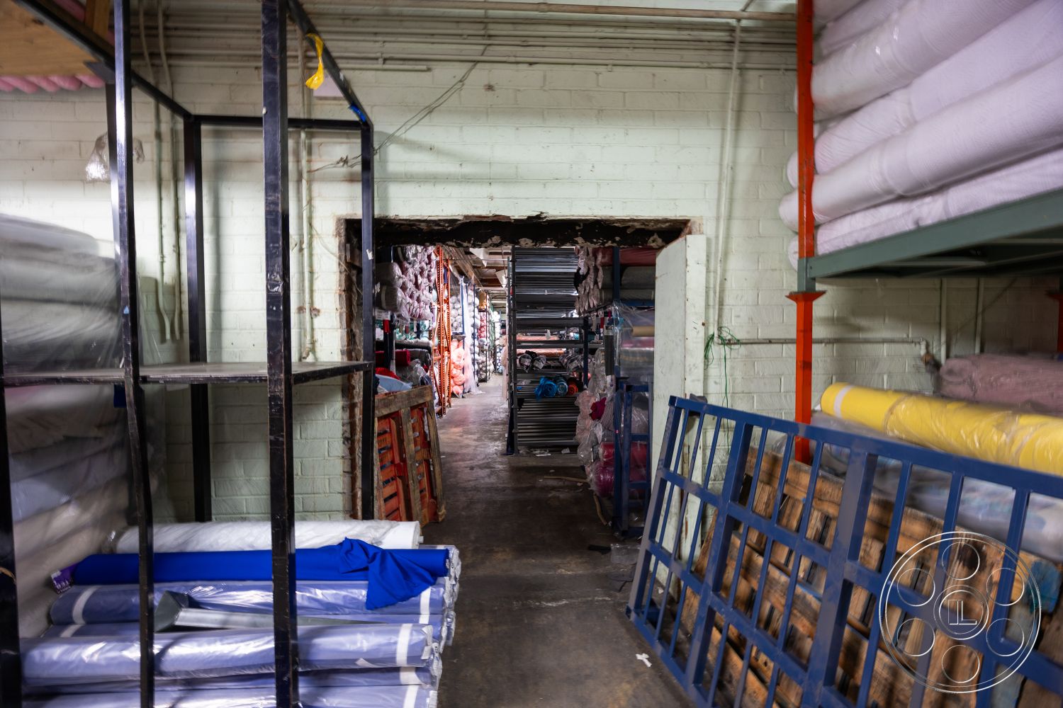 Warehouse 61 - neutral color palette, concrete floor, exposed brick walls, metal shelving units, textured plaster ceiling, stacked pallets, functional work area, open warehouse layout, ambient warehouse lighting, industrial storage space, rolls of fabric stacked, suspended electrical wiring, painted cinder block walls, interior storage facility