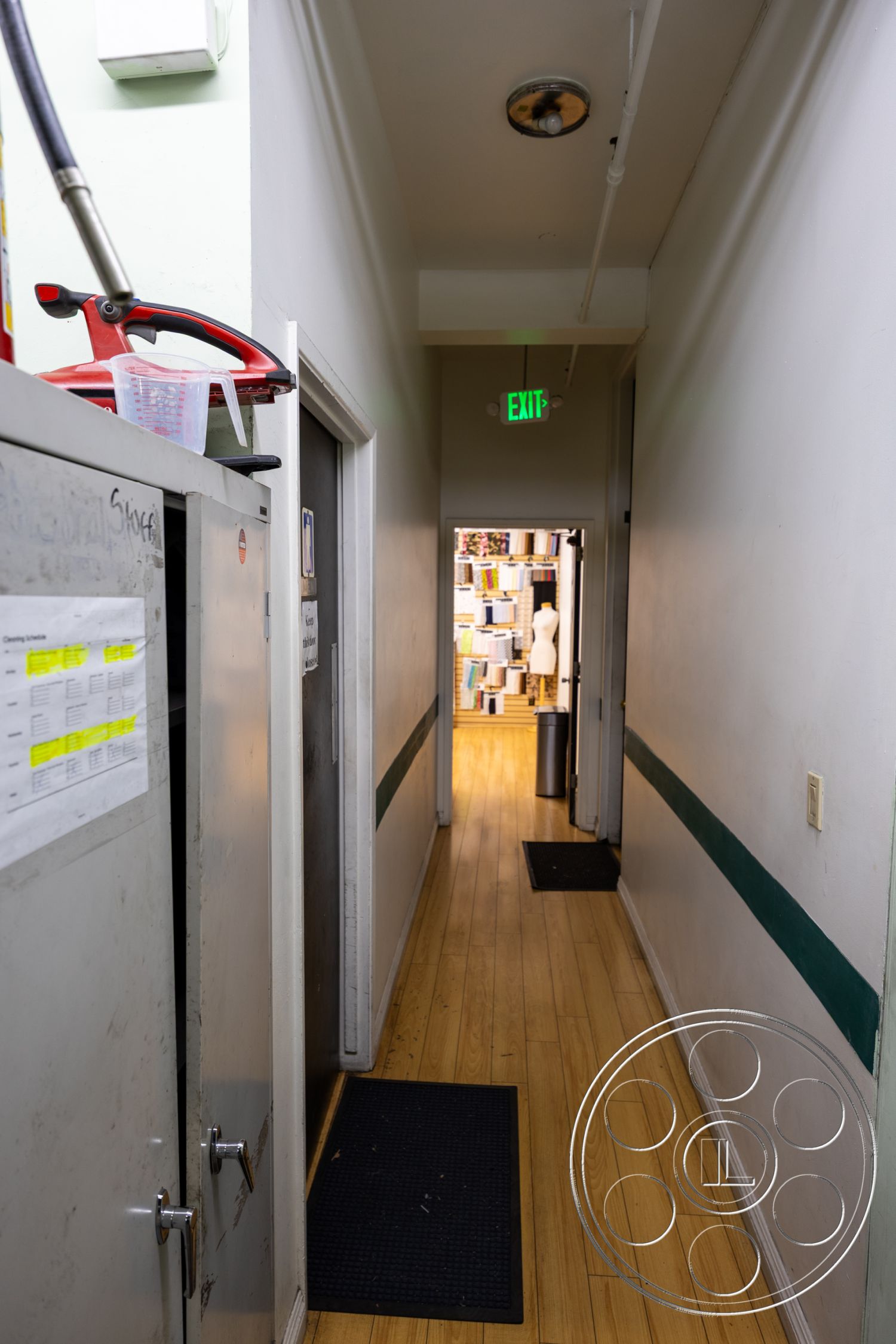 Warehouse 61 - industrial design elements, exit sign, laminate wood flooring, interior hallway, white painted walls, narrow hallway layout, green accent stripe on walls, drop ceiling with light fixtures, metal storage cabinet, black floor mats, closet door, decorative wall treatment, natural light at the end of the hallway, functional workspace access, organized storage area