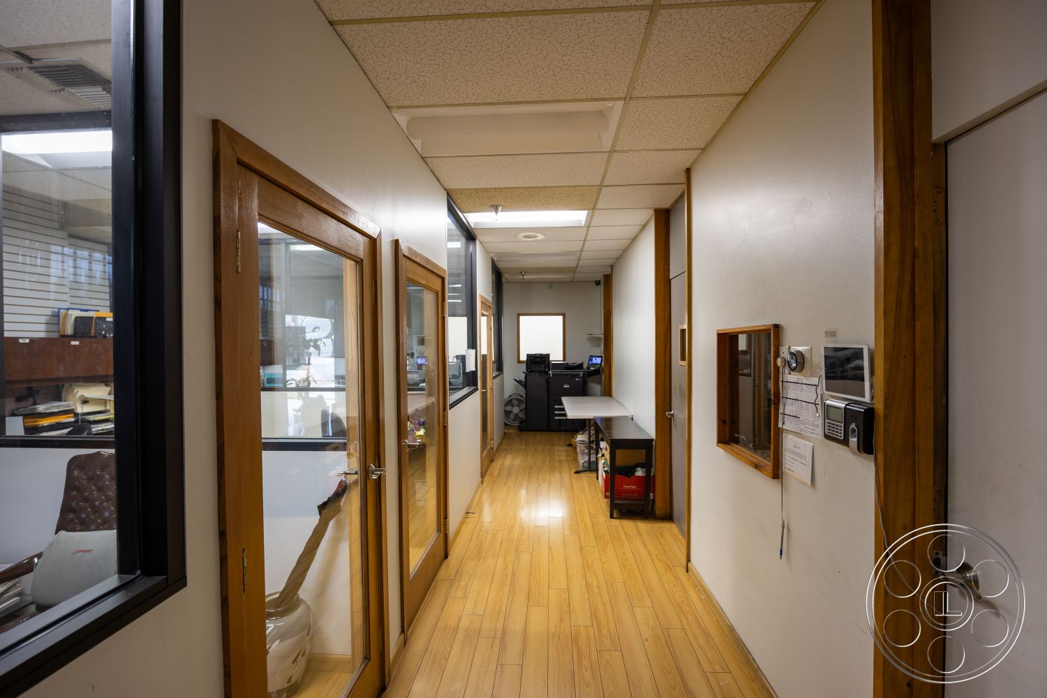 Warehouse 61 - glass partition walls, exposed wood door frames, light wood trim details, textured ceiling tiles, interior corridor with wood laminate flooring, office workspace area with desks, two-panel glass office doors, directory board with metal accents, structured hallway layout, small window overlooking workspace, modern workplace environment
