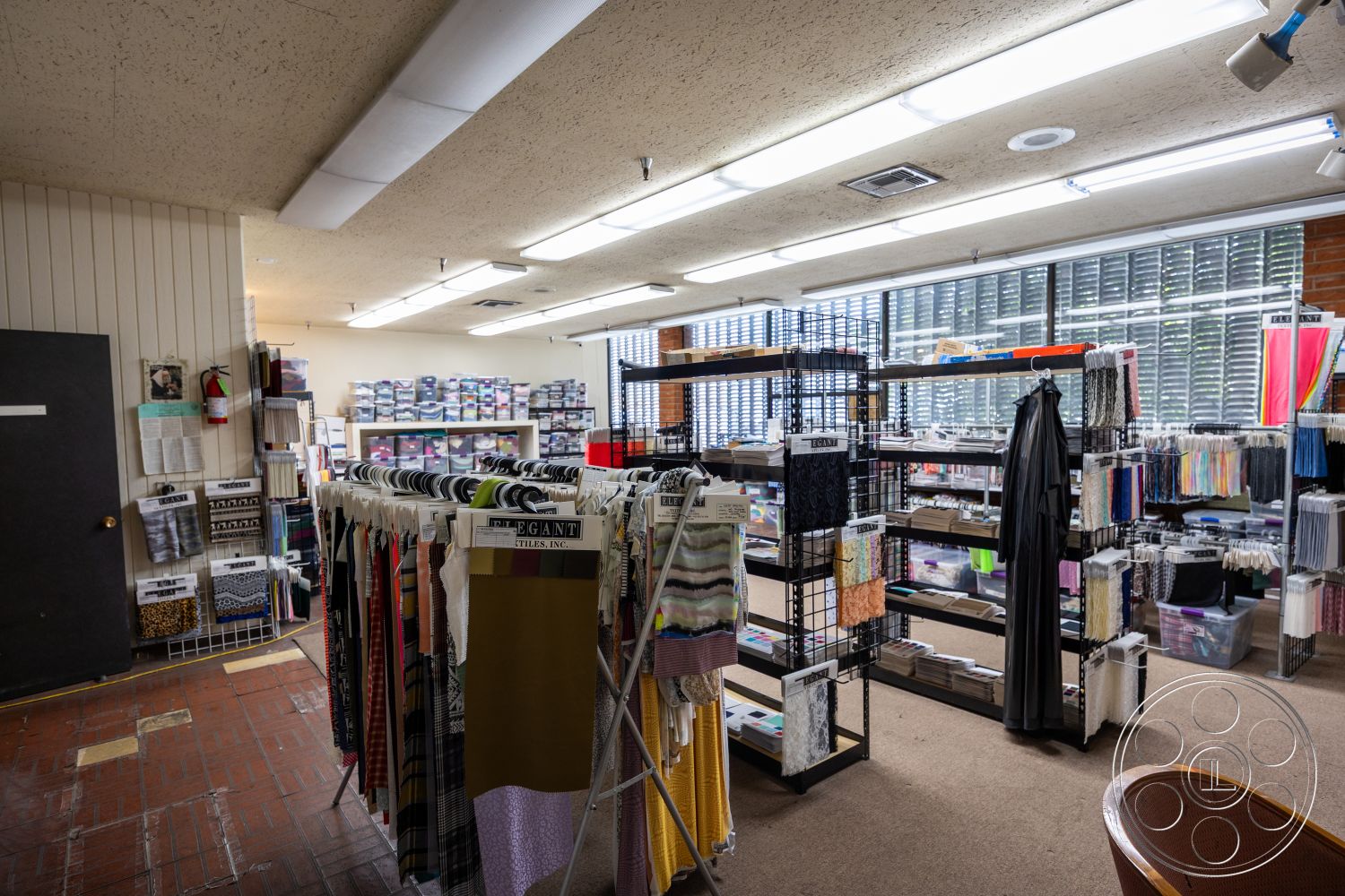 Warehouse 61 - neutral color palette, open floor plan, carpet flooring, decorative wall art, brick accent wall, natural light from large windows, textured plaster walls, wooden shelving units, bright fluorescent lighting, exposed ceiling tiles, ample natural light, organized fabric rolls, interior fabric store, wall-mounted fabric displays, black interior door, wire grid shelving, functional retail layout, storage boxes for fabrics, spacious retail area