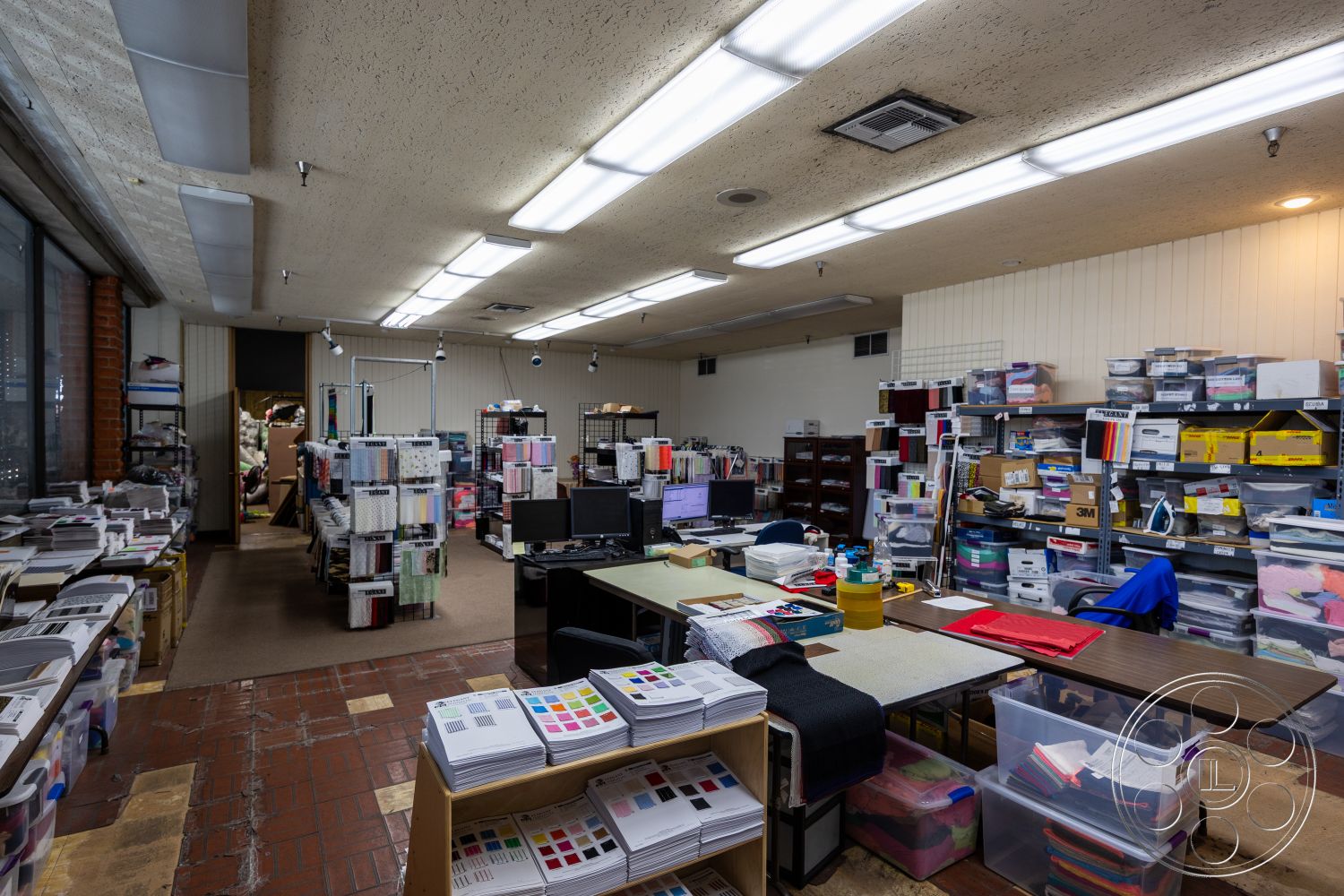 Warehouse 61 - metal shelving units, roll-up garage door, large window view, acoustic ceiling tiles, overhead fluorescent lighting, modern office environment, wood laminate flooring, open workspace layout, exposed brick interior walls, cardboard storage boxes, desktop computer workstation, colorful fabric samples, tile floor pattern, partitioned office space, creative studio setting