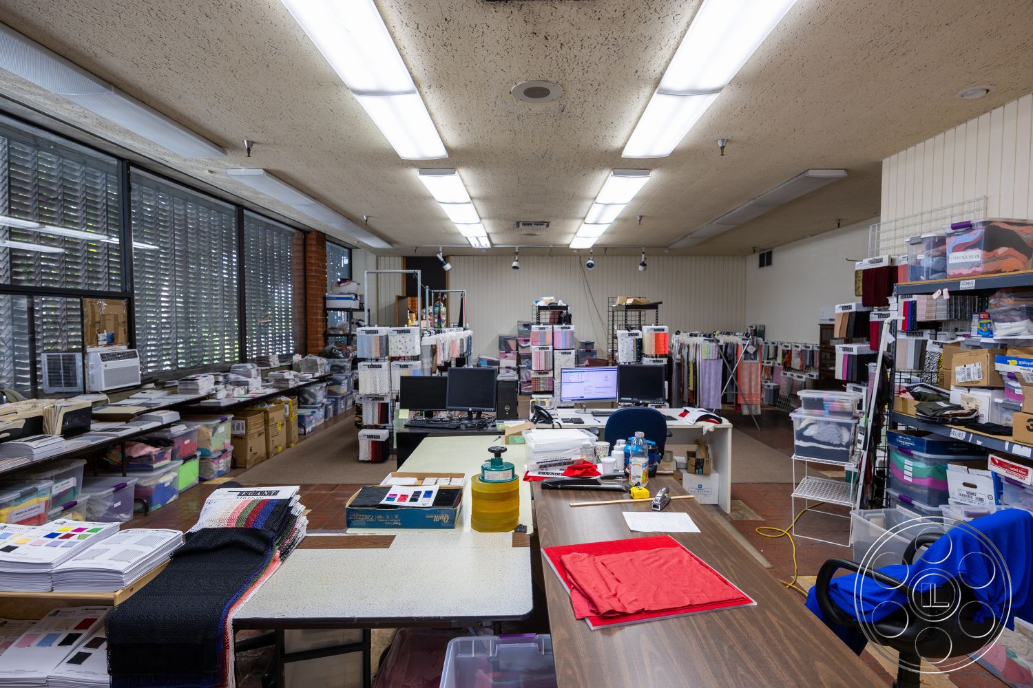 Warehouse 61 - large windows, concrete floor, textured plaster walls, exposed concrete ceiling, industrial light fixtures, metal desk, wooden shelving units, flat roof design, brightly lit interior, commercial office space, functional workstations, open workspace layout, creative workspace environment, laminate countertop, rolling office chairs, fabric swatch display, colorful textile inventory, clean design aesthetics, organized fabric storage, collaborative design area