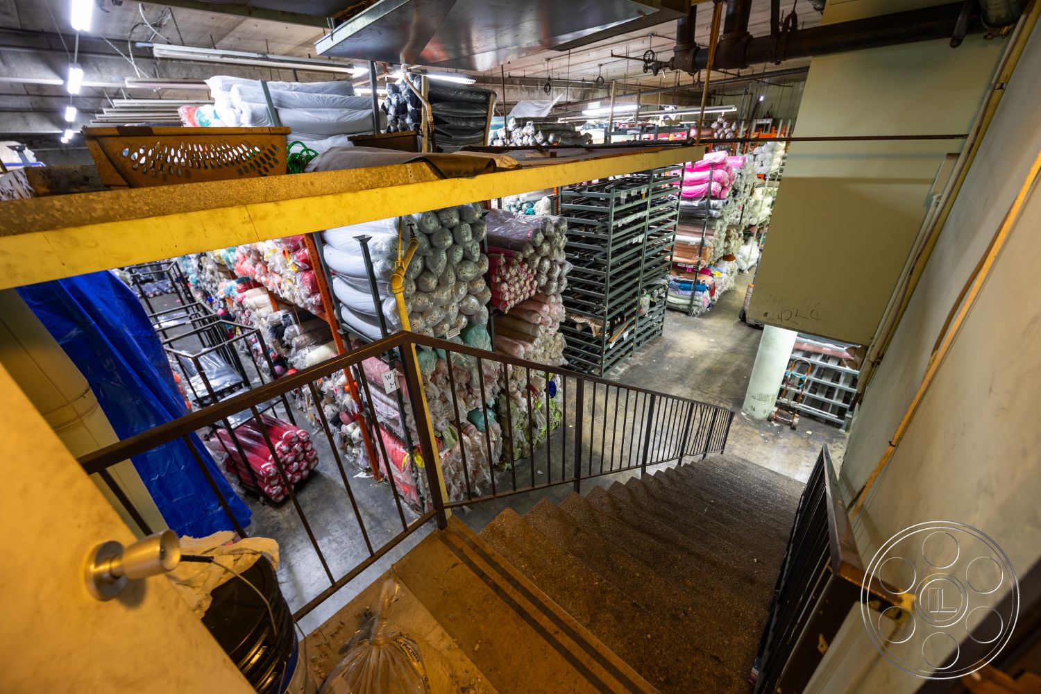 Warehouse 61 - concrete block walls, exposed concrete ceiling, industrial shelving, interior view, overhead fluorescent lighting, metal stair railings, open storage space, concrete staircase steps, industrial interior, textured concrete floor, stacked shelving units, bold color pallets in storage, functional warehouse setting, multicolored fabric rolls, urban basement design, organized inventory display