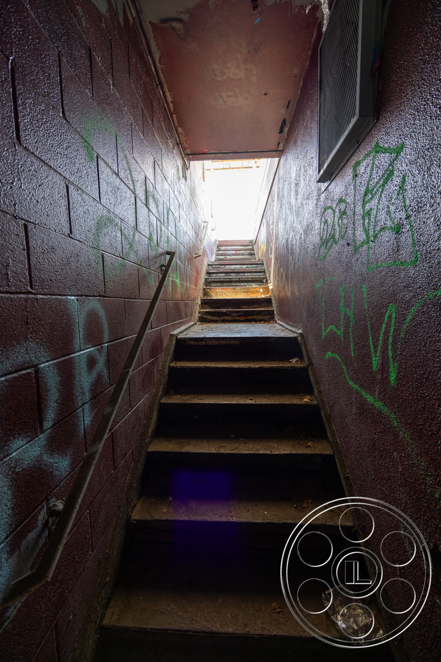Warehouse 61 - exposed concrete block walls, metal stair treads, rounded staircase shape, industrial ceiling with exposed ductwork, faded paint on walls, stair railings with minimalistic design, urban graffiti art details, interior basement stairwell, weathered concrete steps, dimly lit entryway, straight staircase design, crowded with debris and leaves, natural light source from above, functional yet neglected space
