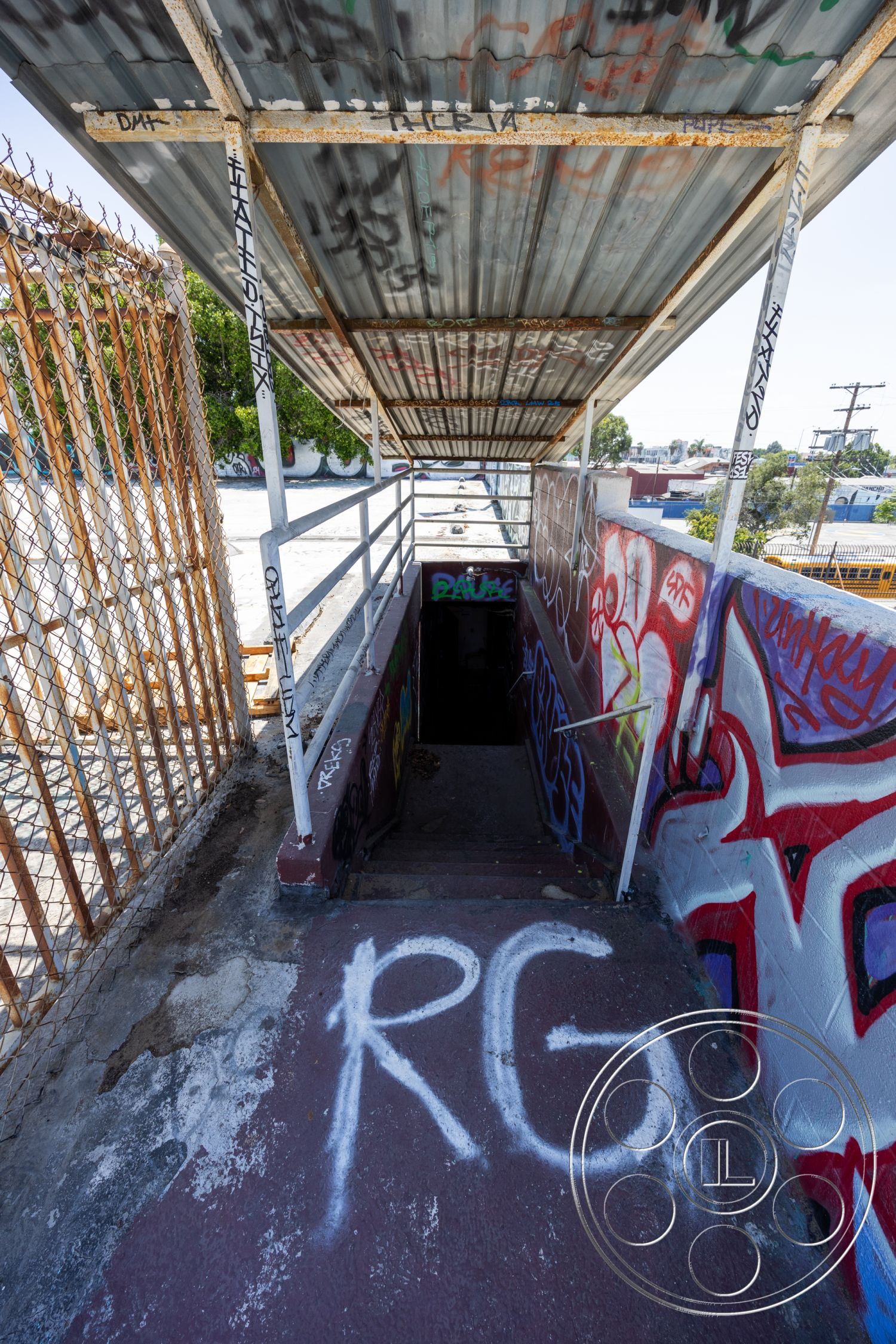 Warehouse 61 - chain link fence, graffiti-covered walls, weathered concrete surface, exterior stairway with concrete steps, metal roof covering, steel railing with graffiti details, dark entrance with no visible lighting, urban alleyway setting, rusted metal structure, open-air entrance with partial shade