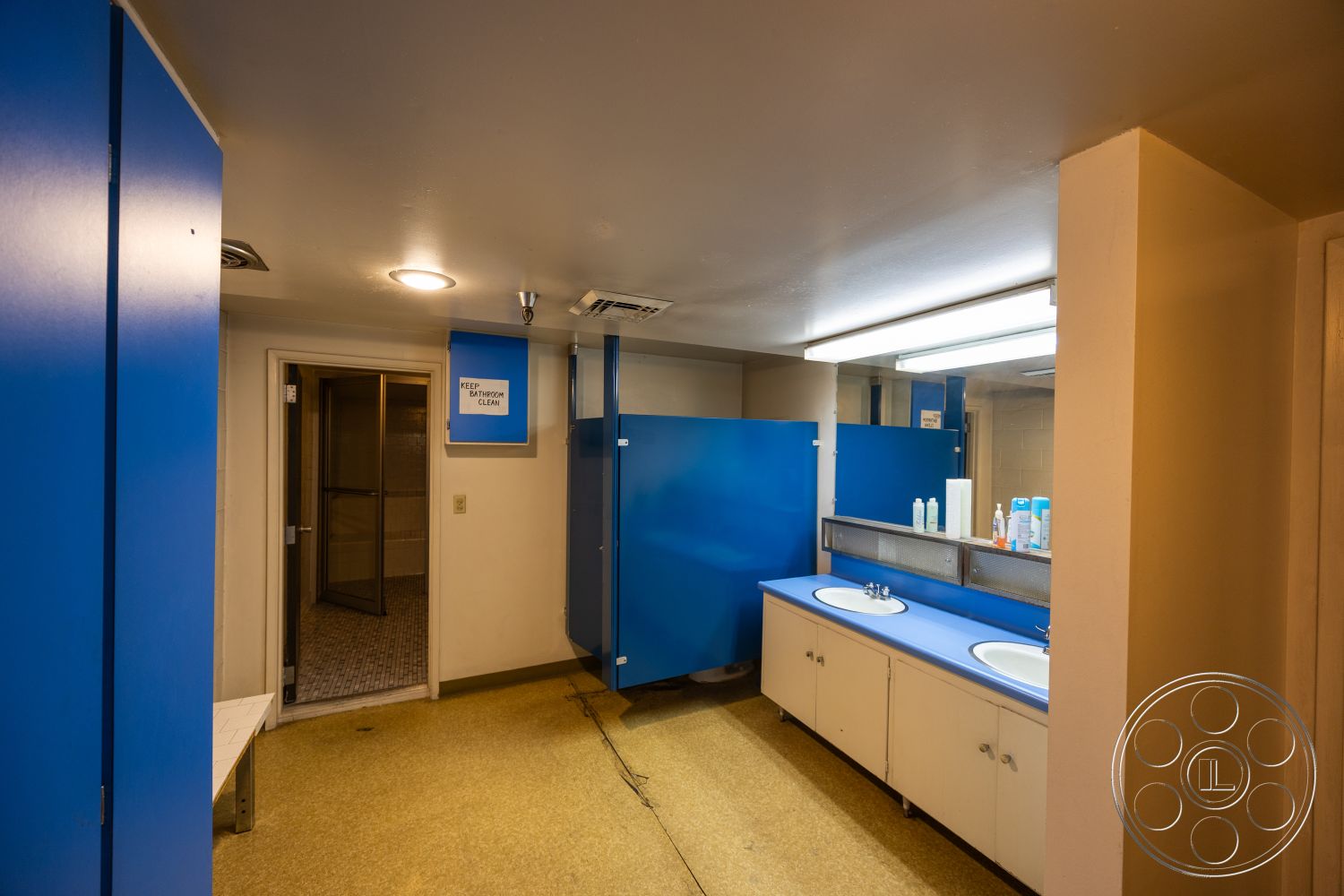 Warehouse 61 - double sink vanity, interior bathroom, white painted walls, fluorescent ceiling lights, grey vinyl flooring, blue laminate cabinetry, white porcelain sinks, mirrored bathroom wall, metal partition walls, enclosed glass shower, accent blue wall panels, access door to shower, functional commercial design, clean and minimalist aesthetic