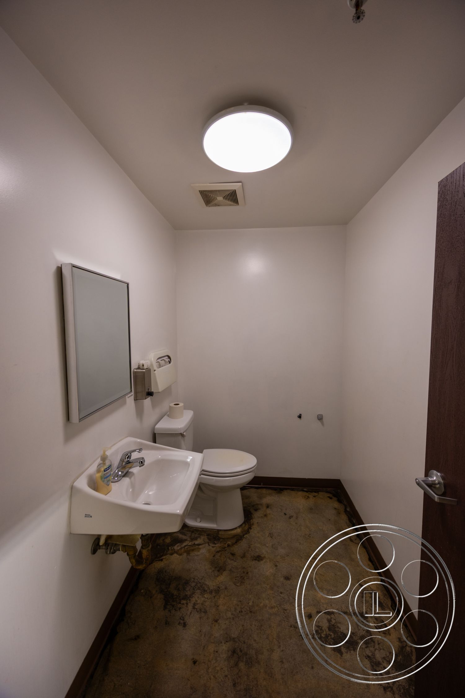 Warehouse 61 - minimalist design, functional layout, concrete flooring, white painted walls, mirror above sink, wall-mounted soap dispenser, exterior bathroom space, modern circular ceiling light, standard toilet, single basin sink with chrome faucet, wooden door trim, basic restroom setting