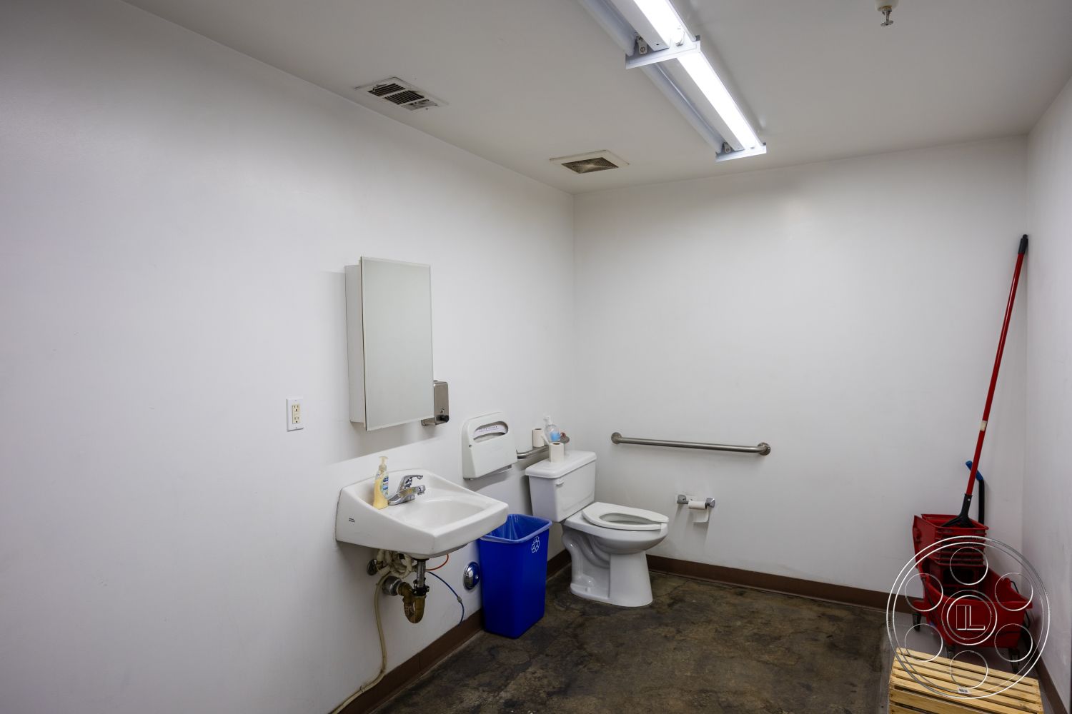 Warehouse 61 - white porcelain toilet, strip lighting on ceiling, accessible restroom features, minimalist bathroom layout, interior bathroom with white painted walls, concrete flooring in commercial bathroom, wall-mounted mirror above sink, stainless steel grab bar for accessibility, utility sink with modern faucet, blue recycling bin in corner, clean, functional design, plumbed for hand dryer, modern restroom signage