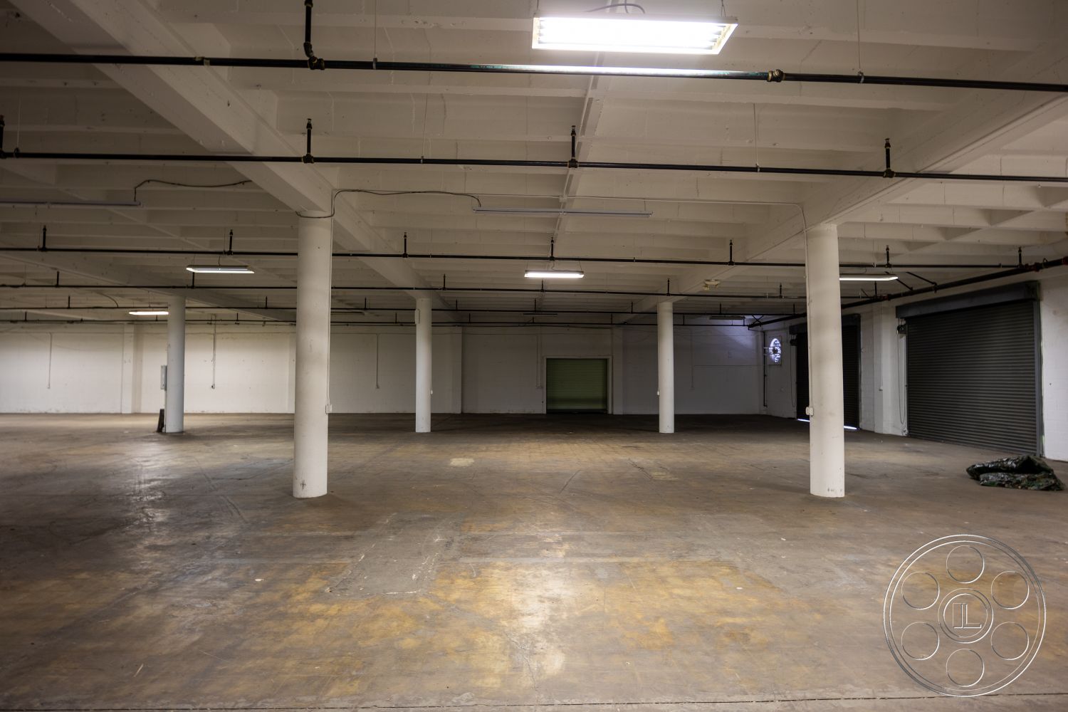 Warehouse 61 - high ceilings, open floor plan, interior space, industrial lighting fixtures, exposed ductwork, roll-up garage door, minimalist architectural style, steel support beams, exposed concrete flooring, white painted concrete walls, concrete column structures, heavy duty electrical wiring, warehouse setting