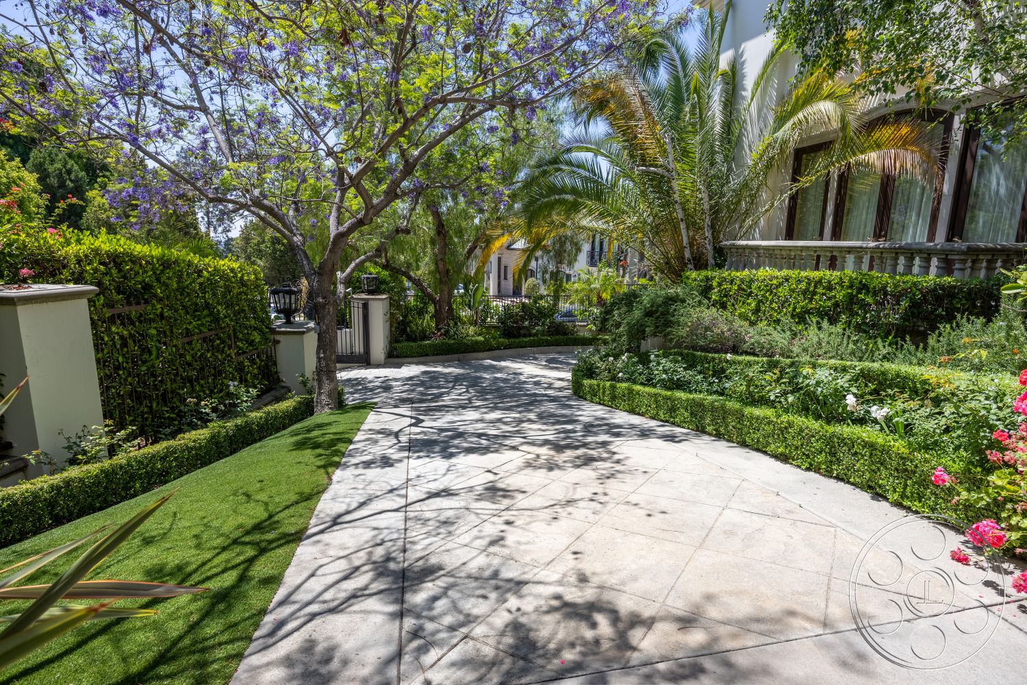 Medit 165 - exterior, curved driveway, tropical palm trees, urban residential setting, mature trees providing shade, concrete driveway pavers, landscape surrounding driveway, formal garden with trimmed hedges, flowering shrubs with pink blooms, lush green lawn with synthetic grass, decorative garden border, fenced perimeter with wrought iron gate