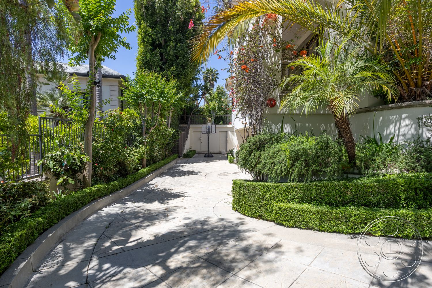 Medit 165 - urban residential setting, exterior concrete driveway, surrounding lush greenery, curved driveway with decorative border, tiled walkway accents, landscaped garden with low hedges, tropical plants with palm trees, modern home exterior with stucco walls, iron fence with decorative gates, exposed aggregate concrete surface, hardscape details with smooth stone, garden patio area with flower beds, softscape elements with flowering vines, sunny environment with clear skies
