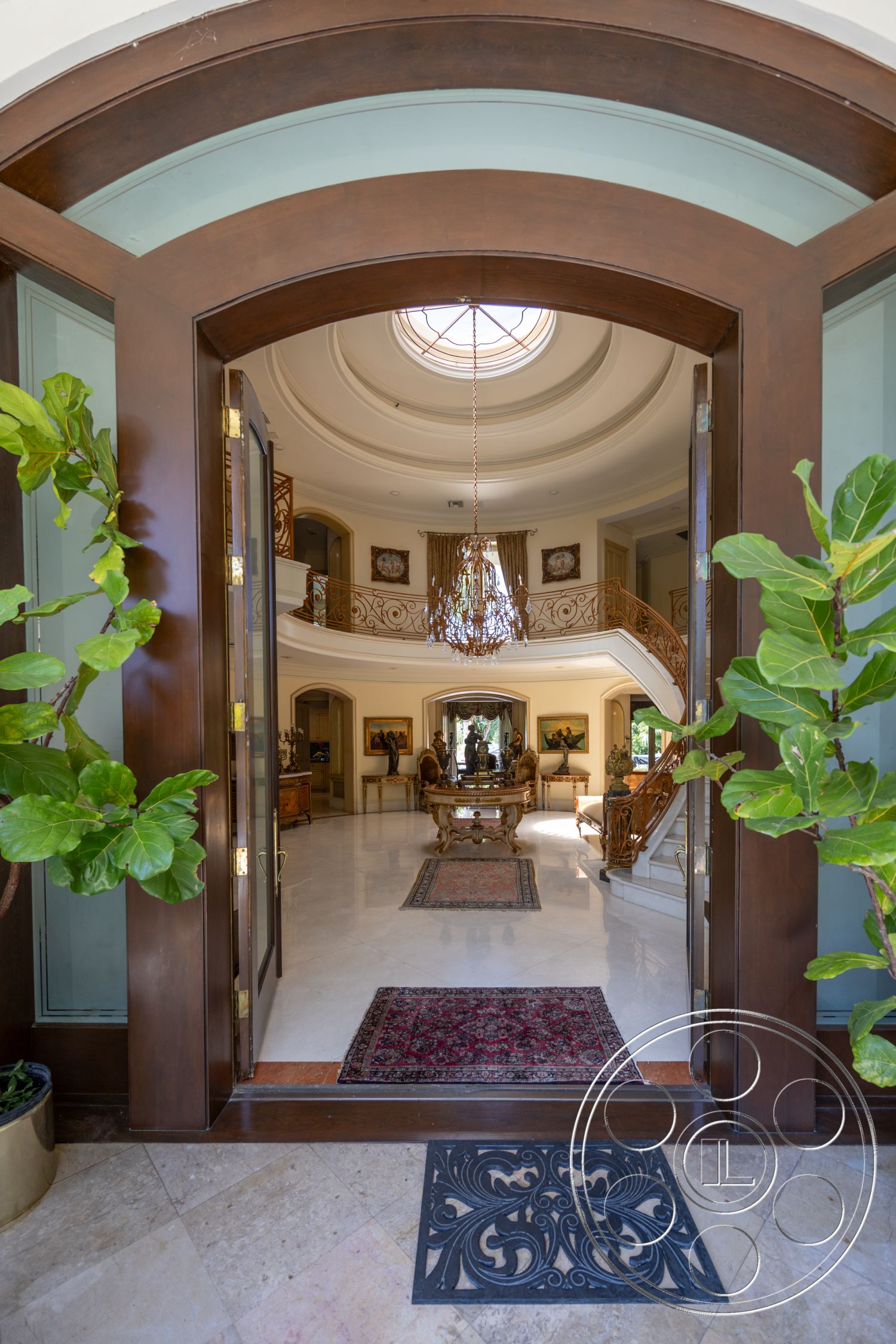 Medit 165 - natural light from large windows, decorative wall sconces, framed artwork on walls, grand entrance foyer interior, arched wooden double doors, marble flooring living room, chandelier lighting feature, crown molding details grand ceiling, curved staircase with iron railings, ornate area rugs on marble floor, plant accents in entryway, textured plaster walls neutral color, high ceilings with decorative medallion, spacious living area with furniture layout, traditional furniture style, exposed wooden elements in design