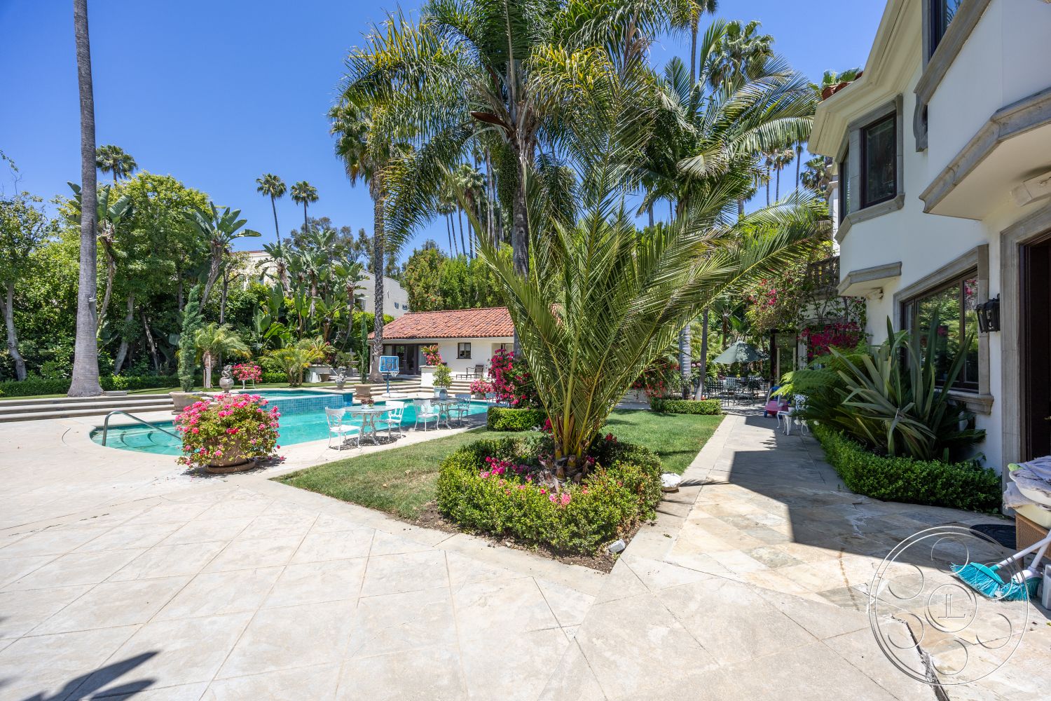 Medit 165 - exterior, landscaped garden, curved pathway, rectangular swimming pool, outdoor dining space, multiple seating areas, stone pool deck, concrete steps leading to pool, green lawn area, lush palm trees, paver patio flooring, surrounded by tropical landscaping, brick outdoor kitchen area, decorative flower beds, attached pool house with red tile roof, interior view of home, large windows with decorative trims, detailed masonry around windows, sunny urban backyard, palm tree framing view