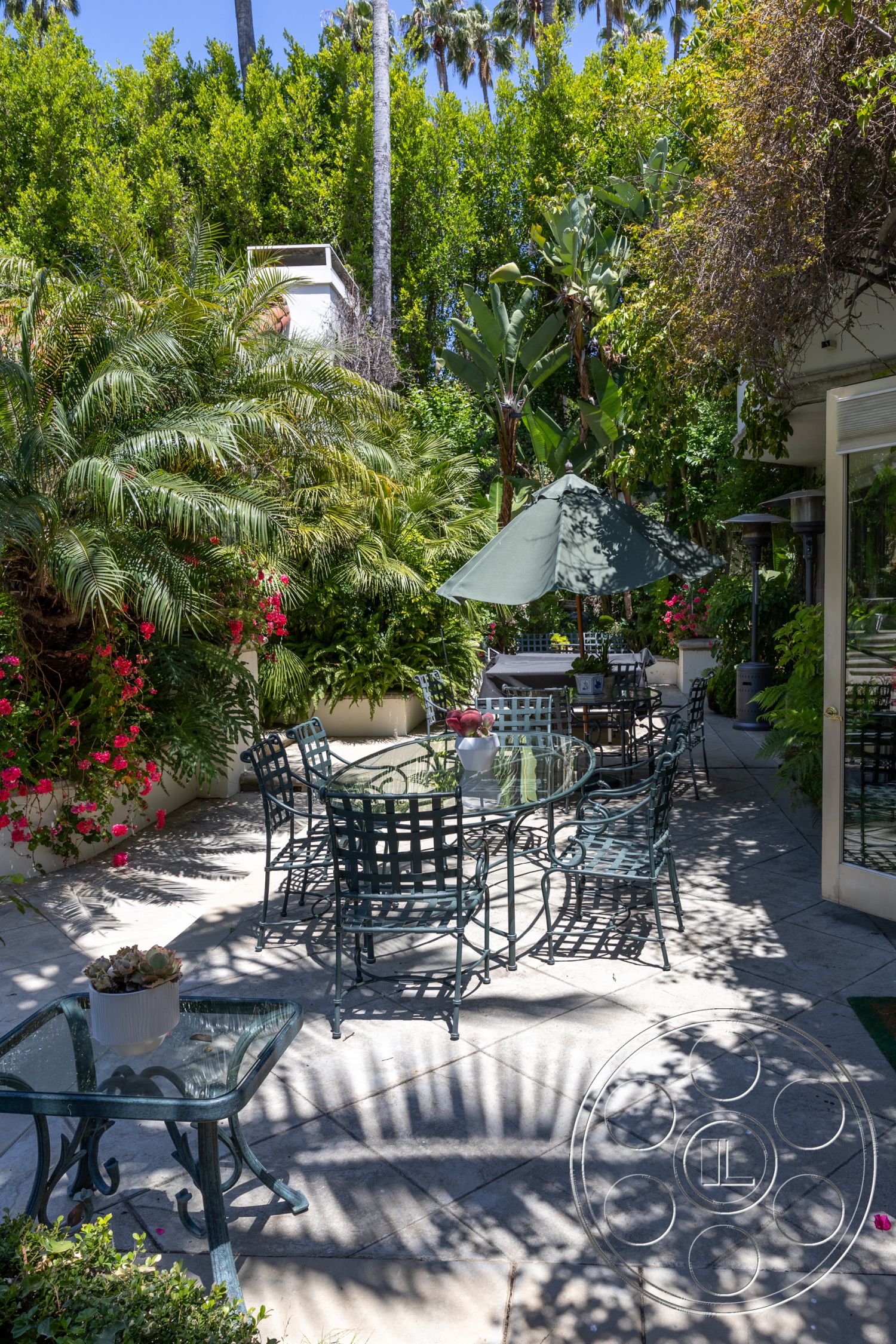 Medit 165 - lush tropical landscaping, tall palm trees, decorative stone wall, metal patio furniture, concrete patio flooring, exterior patio area, glass tabletop, potted succulent plants, green umbrella, vibrant flower garden, shaded outdoor seating, foilage privacy screen, sun dappled shadows, relaxing garden setting, California outdoor living