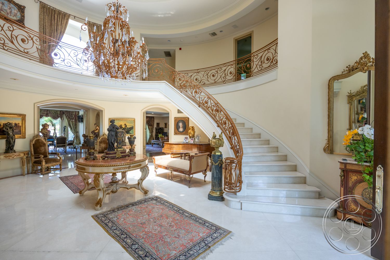 Medit 165 - large arched windows, curved staircase, oriental area rug, coffered ceiling details, high ceiling with crown molding, marble foyer flooring, intricate wrought iron staircase railing, exposed chandelier lighting fixture, gold leaf decor accents, textured cream-painted walls, traditional wood paneling details, ornate gilded mirror, antique artwork framing, sitting area with luxury upholstered chairs, elegant round table, opulent living room setting, interior grand entrance