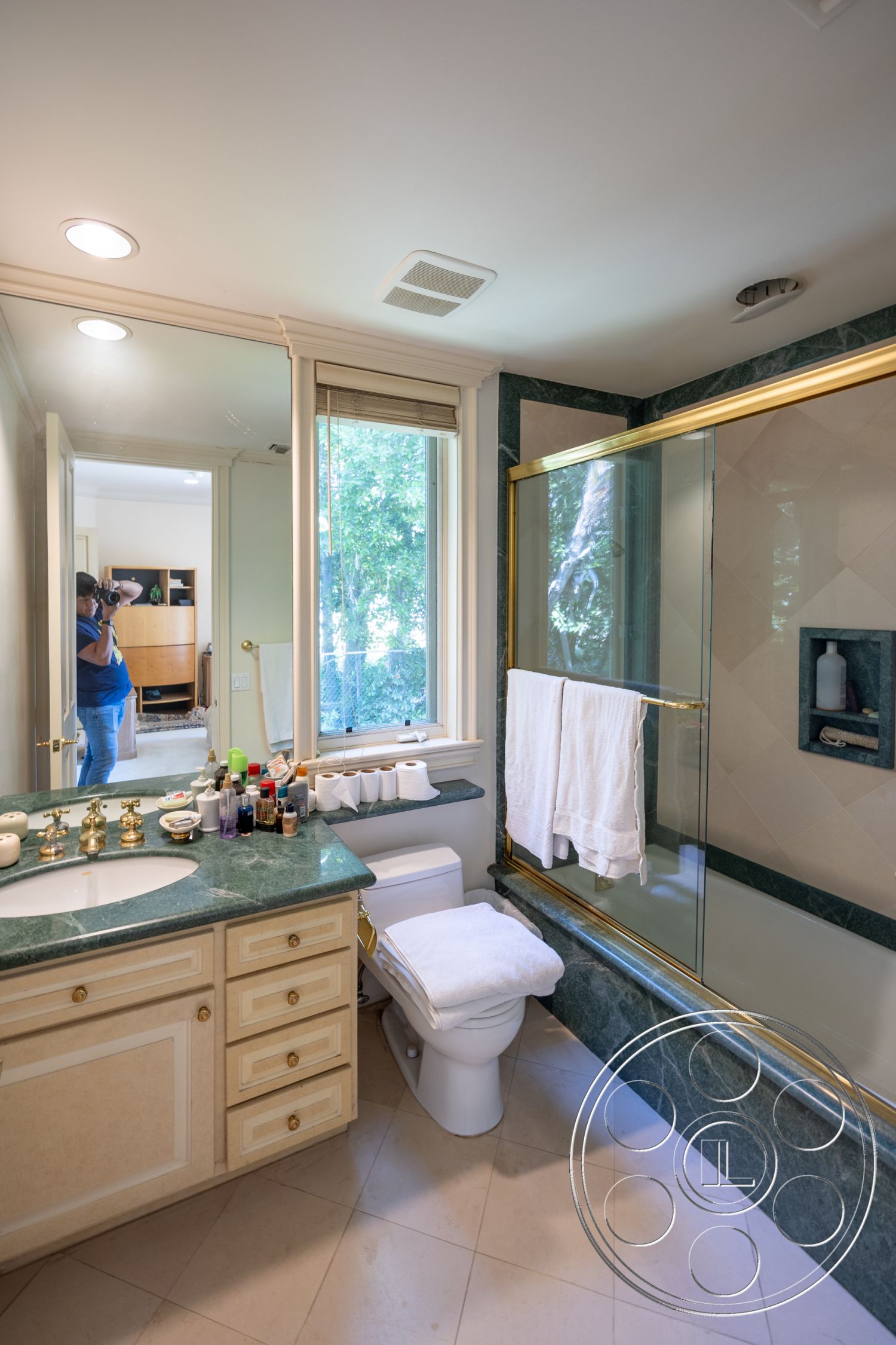 Medit 165 - neutral color palette, natural light from window, tile bathroom flooring, interior bathroom with marble countertop, gold-finished bathroom fixtures, glass shower enclosure with golden trim, green marble bathtub surround, cream-colored cabinetry in bathroom, white towels on towel rack, vanity area with mirror, scenic view through bathroom window, light beige wall paint, classic-style toilet, luxurious bathroom ambiance, accessible storage in bathroom cabinetry, sink area with multiple toiletries