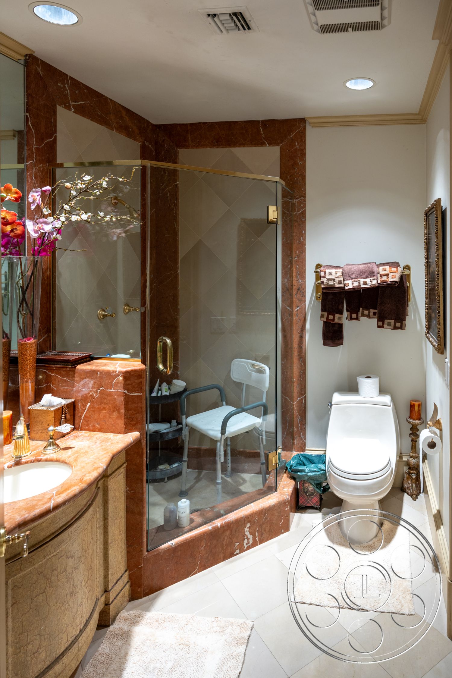 Medit 165 - glass shower enclosure, interior bathroom, marble shower walls, bronze shower hardware, textured beige tile flooring, marble vanity countertop, intricate molding details, decorative gold accents, luxurious towel rack, modern toilet, framed wall mirror, flower arrangement decor, cream-colored area rugs, functional shower chair, golden faucet fixtures, built-in toiletries storage
