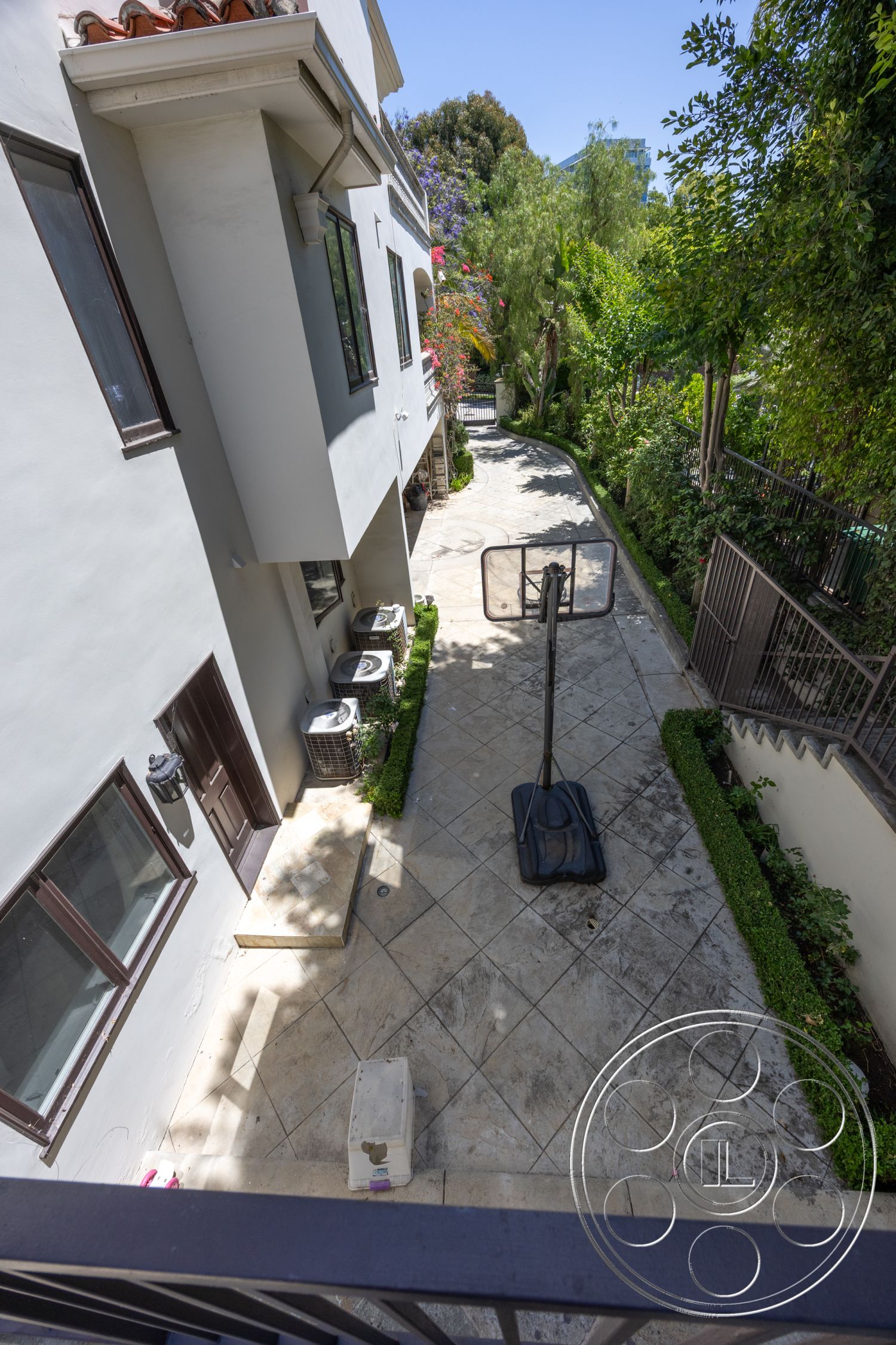 Medit 165 - exterior concrete driveway, exterior lush greenery, exterior concrete patio flooring, exterior stone border landscaping, exterior low-maintenance hedge edging, exterior basketball hoop installation, exterior modern house with stucco walls, exterior double-hung windows with dark frames, exterior tile-paved pathway, exterior air conditioning units, exterior residential setting with garden, exterior multi-level property, exterior urban environment with nearby buildings, exterior smooth wall finish, exterior decorative entryway