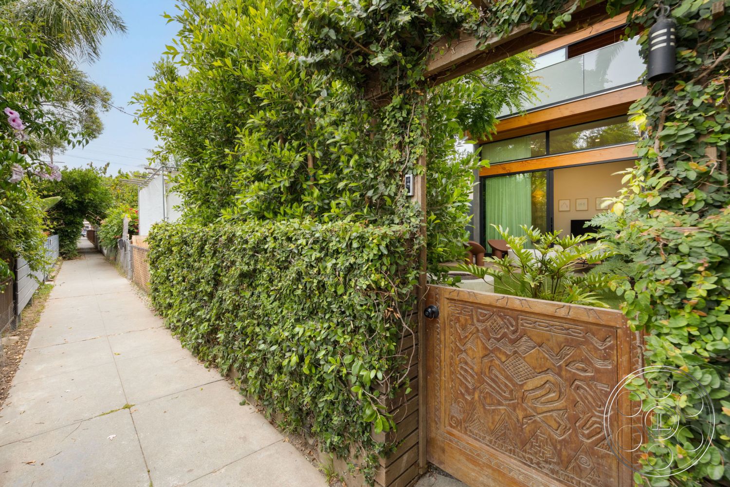 Modern 508 - natural light-filled living space, exterior wooden gate with intricate carvings, lush green hedge landscaping, sidewalk pathway, modern glass windows, wooden siding façade, contemporary residential design, tropical garden atmosphere, interior view through large windows, mixed materials in entrance area, urban garden entrance, decorative wooden gate details, surrounding lush foliage