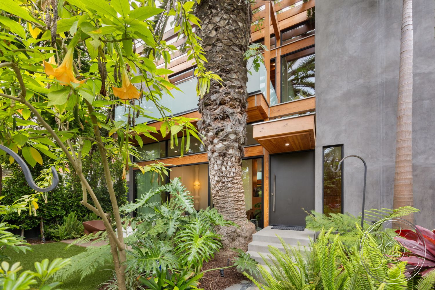 Modern 508 - low-maintenance landscaping, exposed concrete walls, contemporary architecture, staircase with metal railing, landscaped garden setting, wooden entry door, framed glass windows, wooden roof overhang, flat-roofed modern exterior, lush tropical vegetation, concrete steps leading to entry, multicolored foliage plants, urban outdoor living space, natural stone planter, interior lighting features visible through windows, modern design aesthetics, raw material finishes, eco-friendly construction materials, dense palm trees in garden, smooth concrete path
