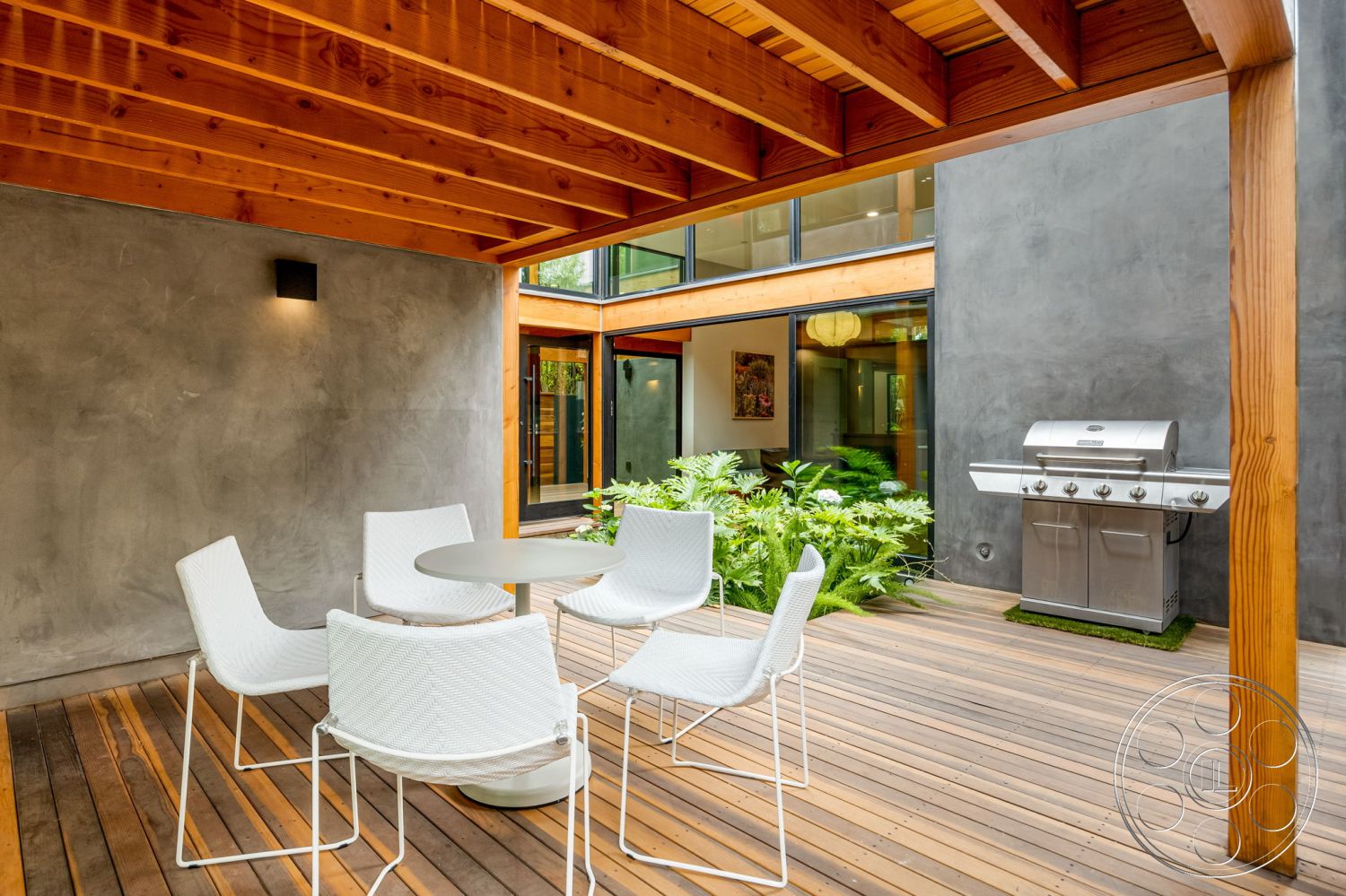 Modern 508 - natural light from large windows, interior glass wall partitions, exposed wooden ceiling beams, exterior patio with hardwood decking, modern white outdoor dining chairs, round outdoor dining table, smooth concrete wall finish, stainless steel outdoor grill, lush green plants surrounding patio, minimalist outdoor furniture design