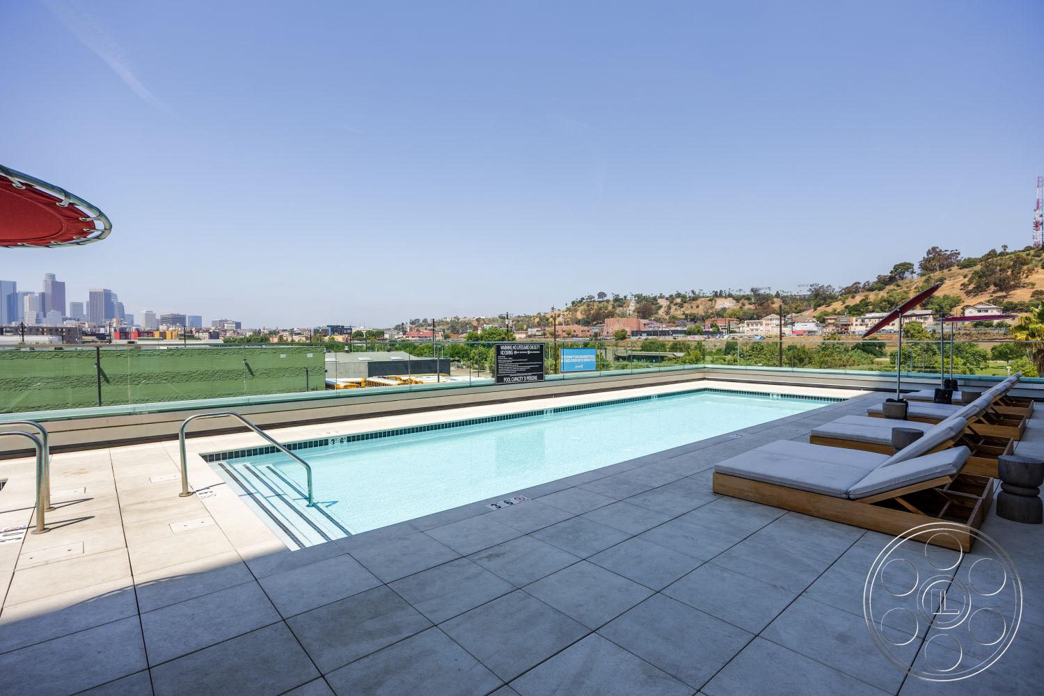 Apartment 55 - glass railing, rectangular swimming pool, modern lounge chairs, concrete patio flooring, urban skyline backdrop, exterior pool area, ceramic tile pool deck, landscaped hillside view, outdoor sunshade umbrellas, textured concrete surround, luxury rooftop deck, city view terrace, minimalist architecture details