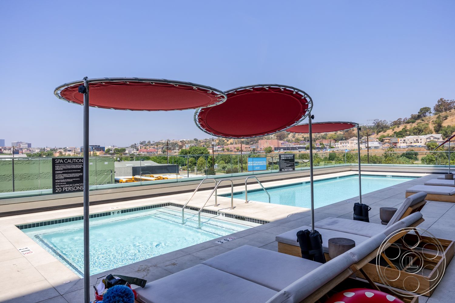 Apartment 55 - glass railing, landscape view, rectangular swimming pool, concrete pool deck, modern outdoor seating, urban skyline backdrop, exterior pool area, red fabric pool umbrellas, stone lounge area, sunny rooftop setting