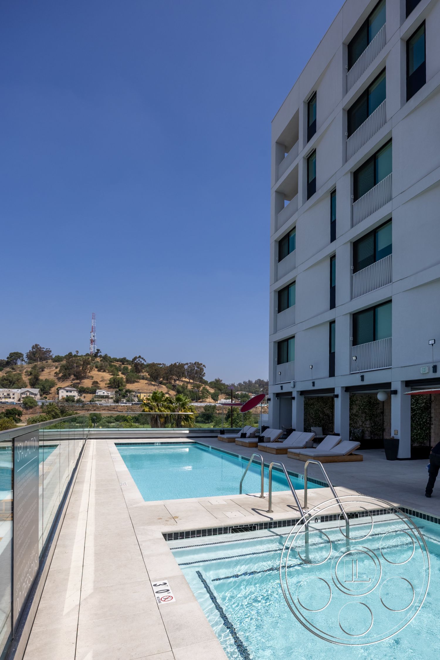 Apartment 55 - rectangular swimming pool, outdoor lounge area, poolside lounge chairs, urban hillside location, concrete patio flooring, glass window treatments, exterior concrete pool deck, glass railing balcony, sunny outdoor environment, white stucco exterior walls, modern building facade, exterior landscaping with palm trees, landscaped hillside setting, tile pool border, multi-story contemporary hotel design