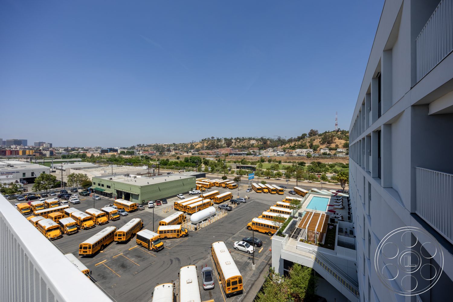 Apartment 55 - rectangular swimming pool, urban setting, industrial architecture, landscaped green area, asphalt parking lot, exterior concrete walkway, concrete exterior walls, metal fence railings, flat rooftop terrace, downtown view, school bus fleet, vibrant yellow school buses, modern commercial building, multi-story parking structure, surrounding hillside landscape