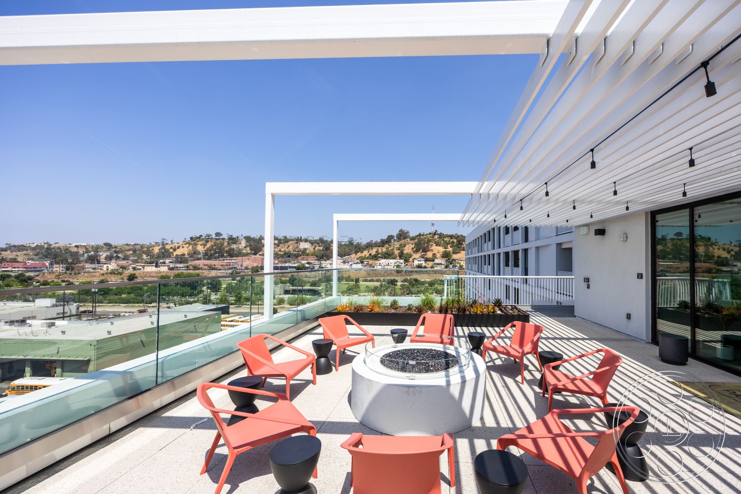 Apartment 55 - minimalist design elements, concrete patio flooring, contemporary outdoor seating area, glass balcony railing, urban skyline backdrop, exterior rooftop patio, modern circular fire pit, steel pergola structure, red molded plastic outdoor chairs, landscape view setting, horizontal slat design, sunny rooftop terrace, textured outdoor flooring, stylish outdoor living space, environmental landscape with hills