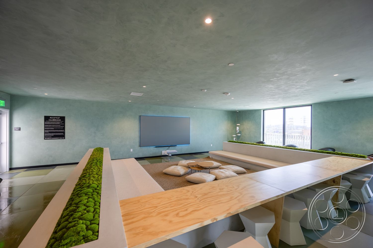Apartment 55 - modern pendant lighting, interior lounge space, contemporary furniture arrangement, concrete floor with geometric patterns, green textured plaster walls, plywood communal seating area, natural moss accent decor, floor-to-ceiling windows with urban view, soft cushioned seating, yoga-style floor cushions, open-plan design with minimalistic features, greenery integrated into furniture design, built-in wooden table surface, urban rooftop clubhouse, light fixtures embedded in ceiling