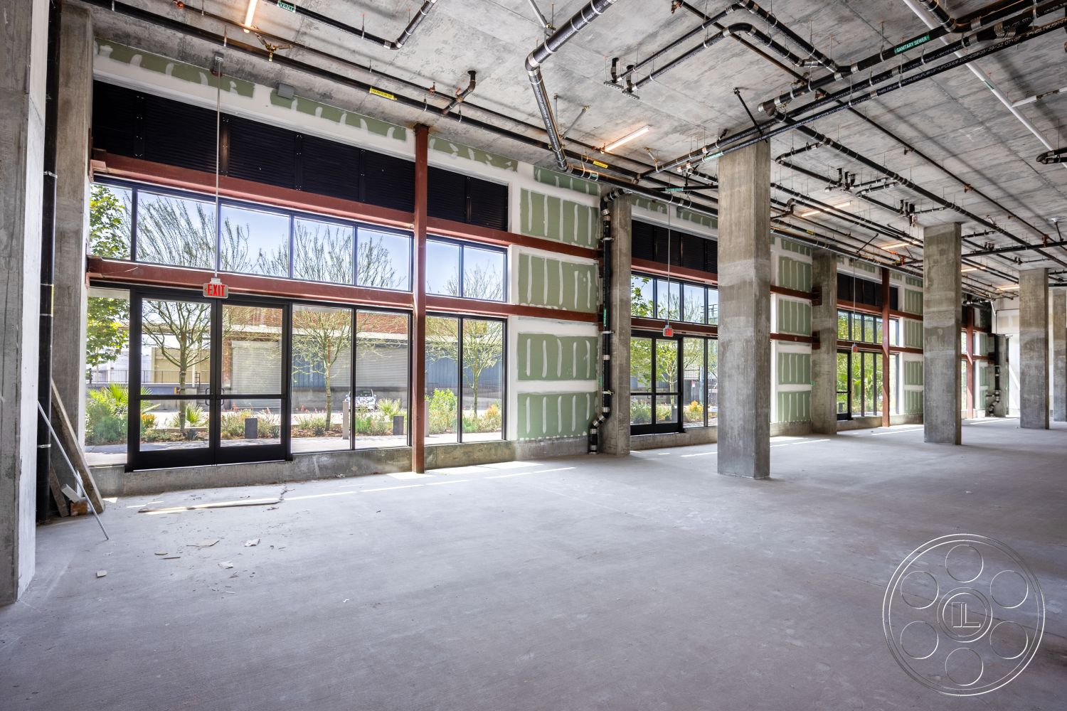 Apartment 55 - open floor plan, concrete flooring, exposed concrete ceiling, drywall partitions, natural light through large windows, open layout design, steel support beams, glass storefront windows, urban commercial space, industrial-style interior, large unobstructed spaces, columns with concrete finish, modern structural elements, interior under construction, pipe infrastructure overhead