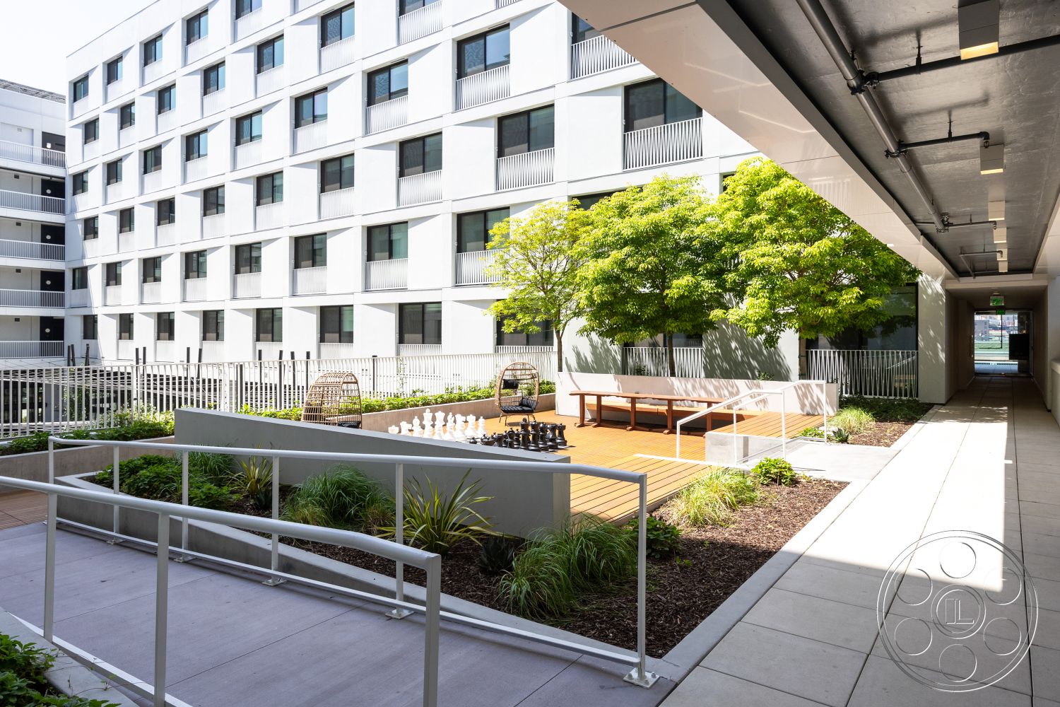 Apartment 55 - contemporary architectural style, urban courtyard setting, tile patio flooring, exterior courtyard with landscaping, concrete walkway with accessibility ramp, wooden bench seating area, chessboard styled outdoor chess set, surrounding greenery with shrubs and trees, modern multi-story residential building, glass railings on balconies, rectangular wooden deck surface, open outdoor area for social gatherings, metal fencing for security, brightly lit walkways with overhead lighting, lawn area with low-maintenance plants